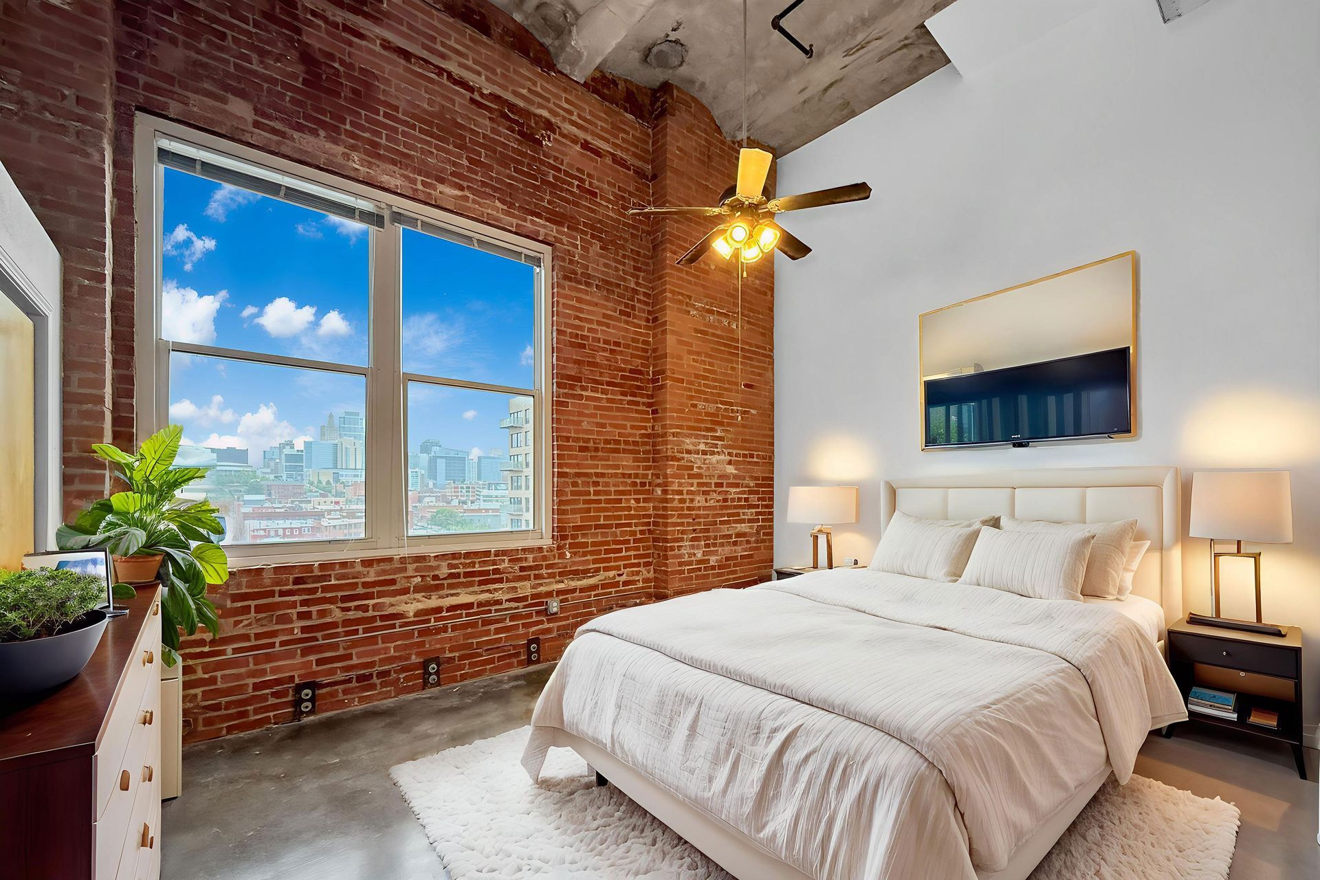 A bedroom with a queen bed, white bedding, exposed brick wall, large window with a city view, and concrete floors.