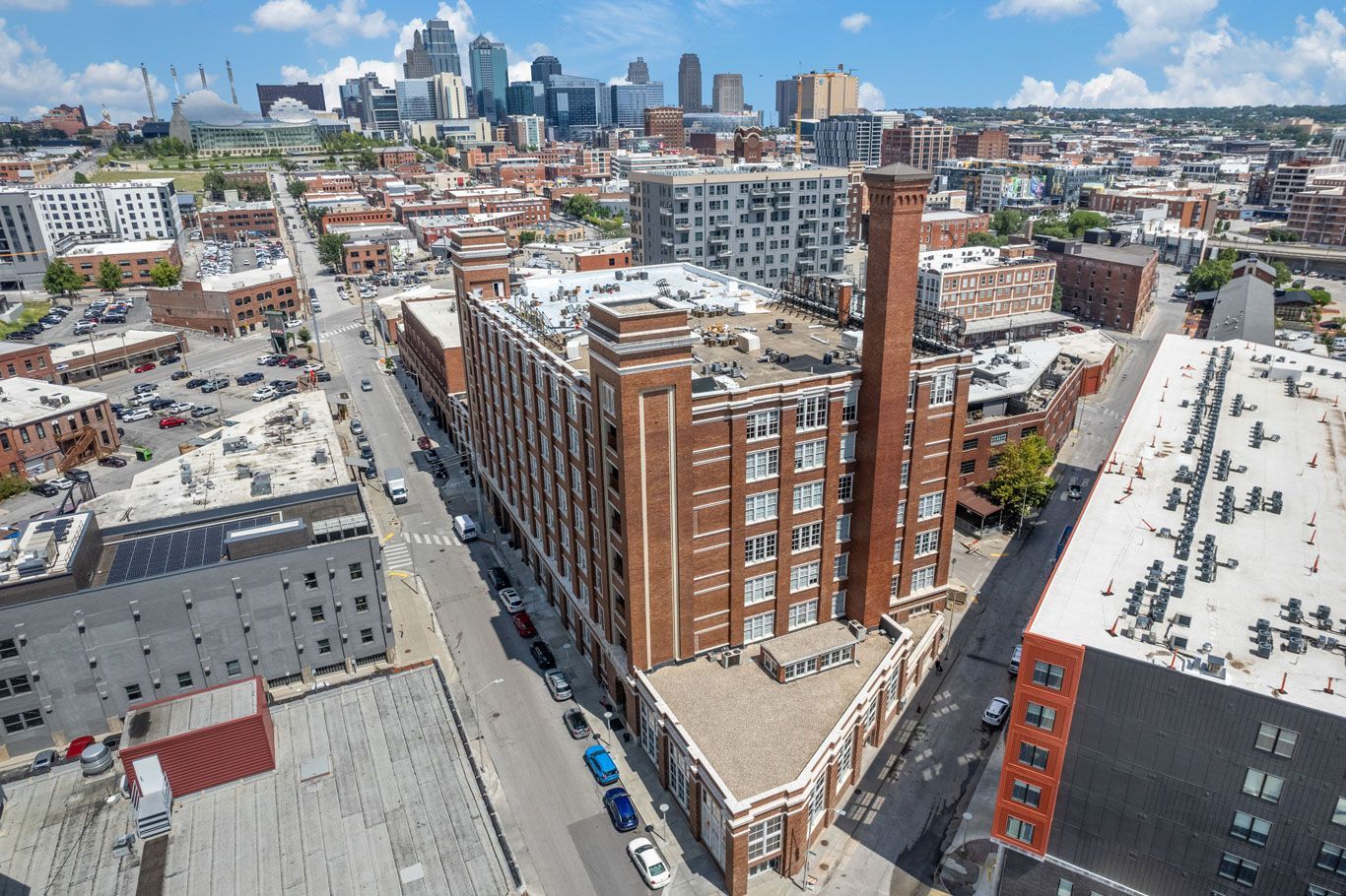 Aerial view of a historic brick building with a tall chimney, situated in a city street grid with a skyline in the back.