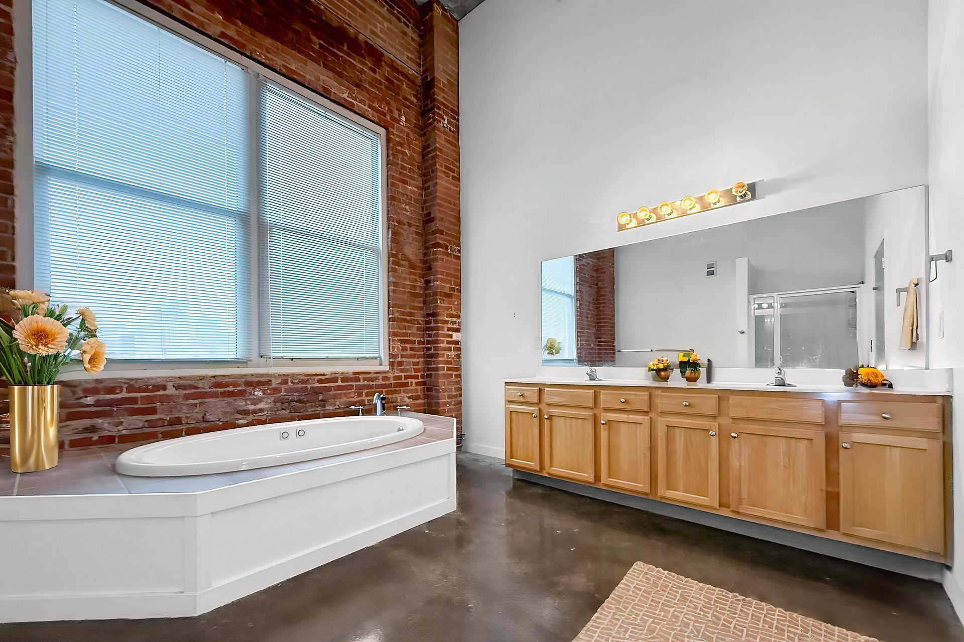 A bathroom with a soaking tub by a large window, exposed brick wall, and a long wooden double vanity under a wide mirror.