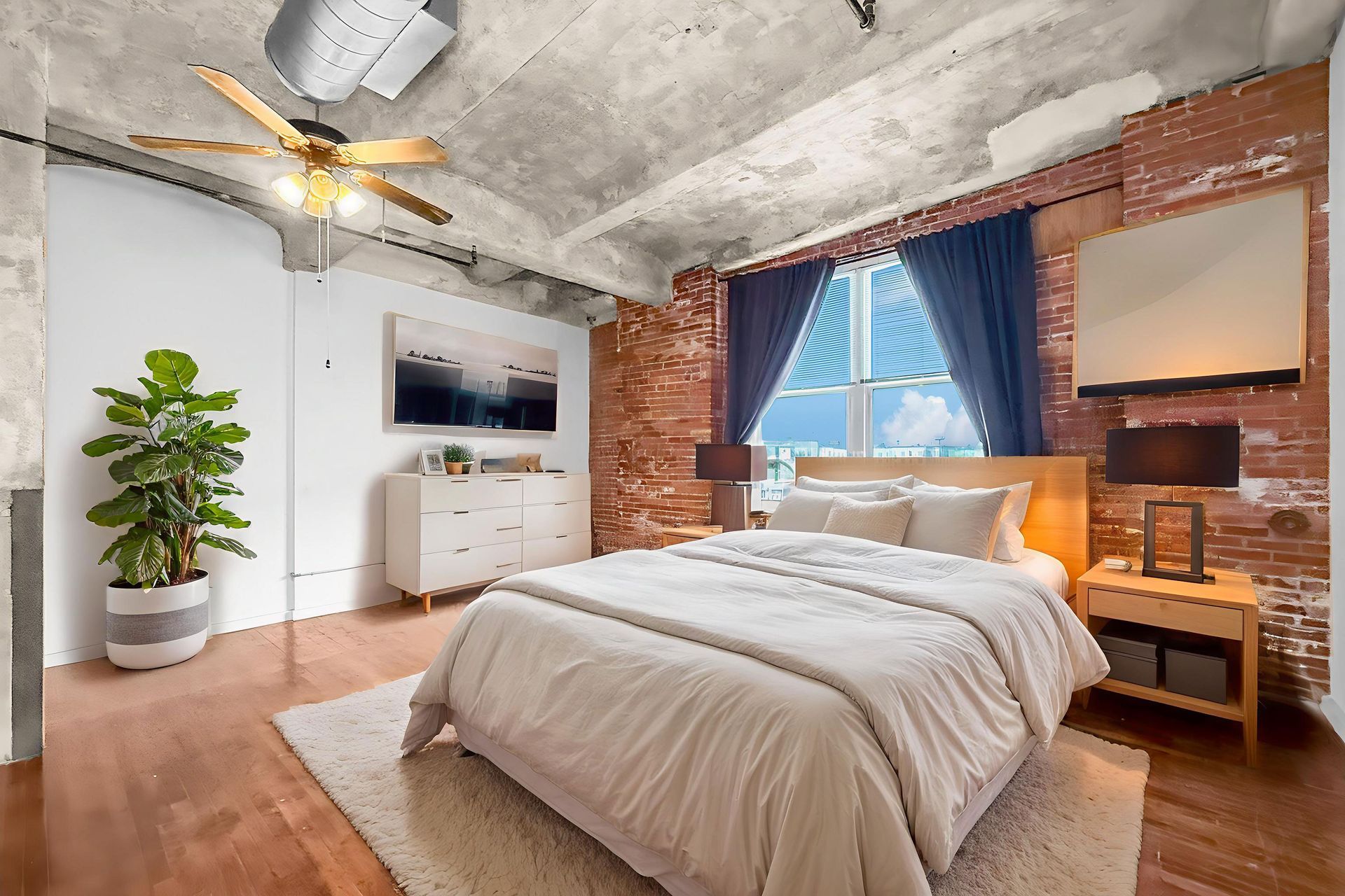 A bedroom with a bed, white bedding, nightstand, dresser, and a large plant against an exposed brick wall and concrete ceiling.