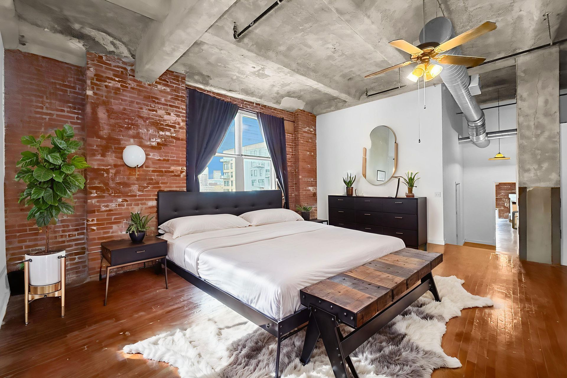 A loft bedroom with an exposed brick wall, white bedding on a dark bed frame, hardwood floors, and a concrete ceiling.