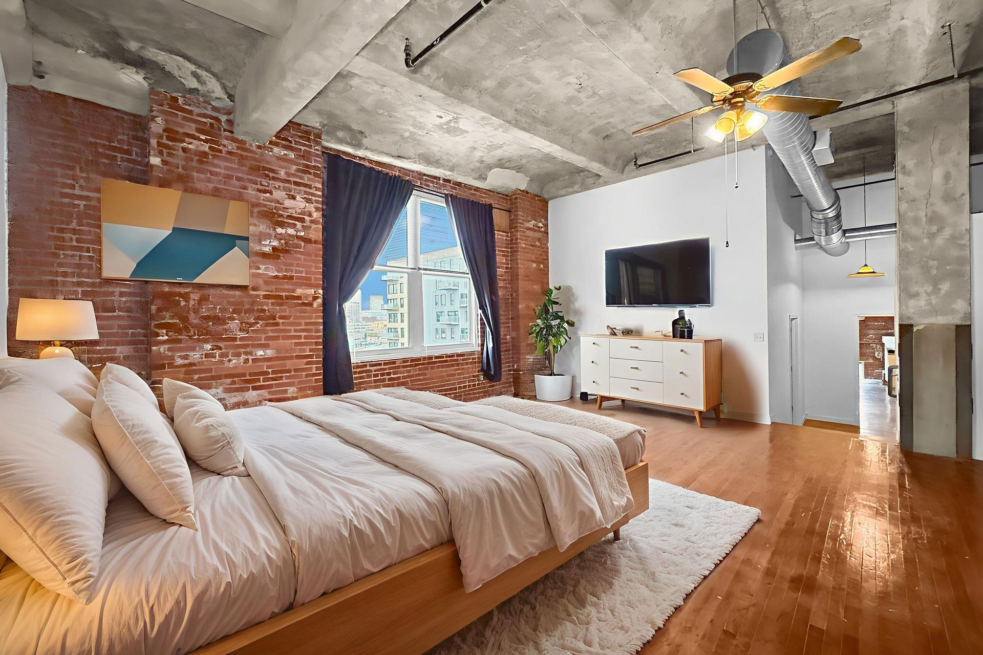 A modern bedroom with a brick wall, concrete ceiling, wooden bed with neutral bedding, and a flat-screen TV.