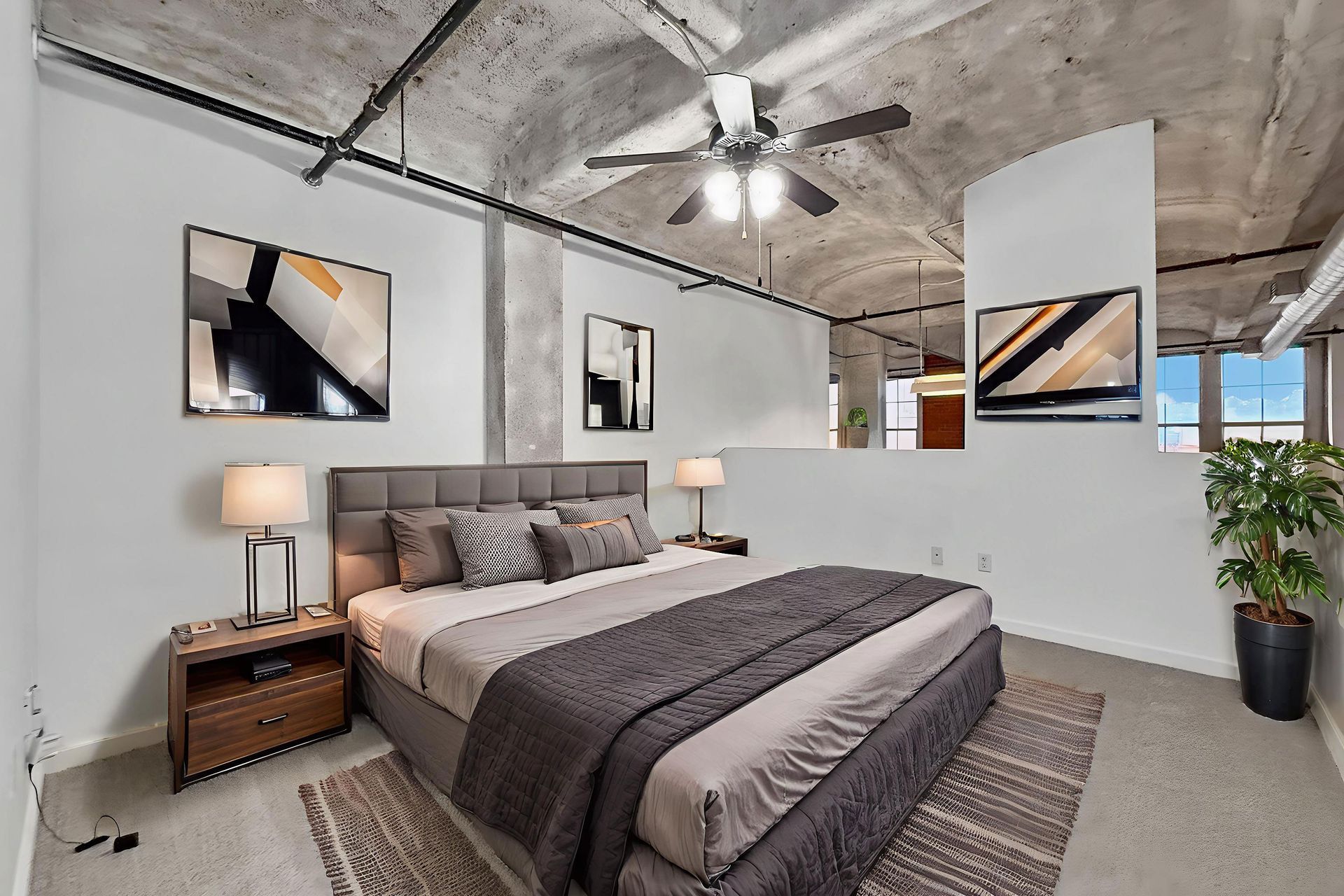 A bedroom with a king-sized bed, exposed concrete ceiling, ceiling fan, neutral walls, and a potted plant in the corner.