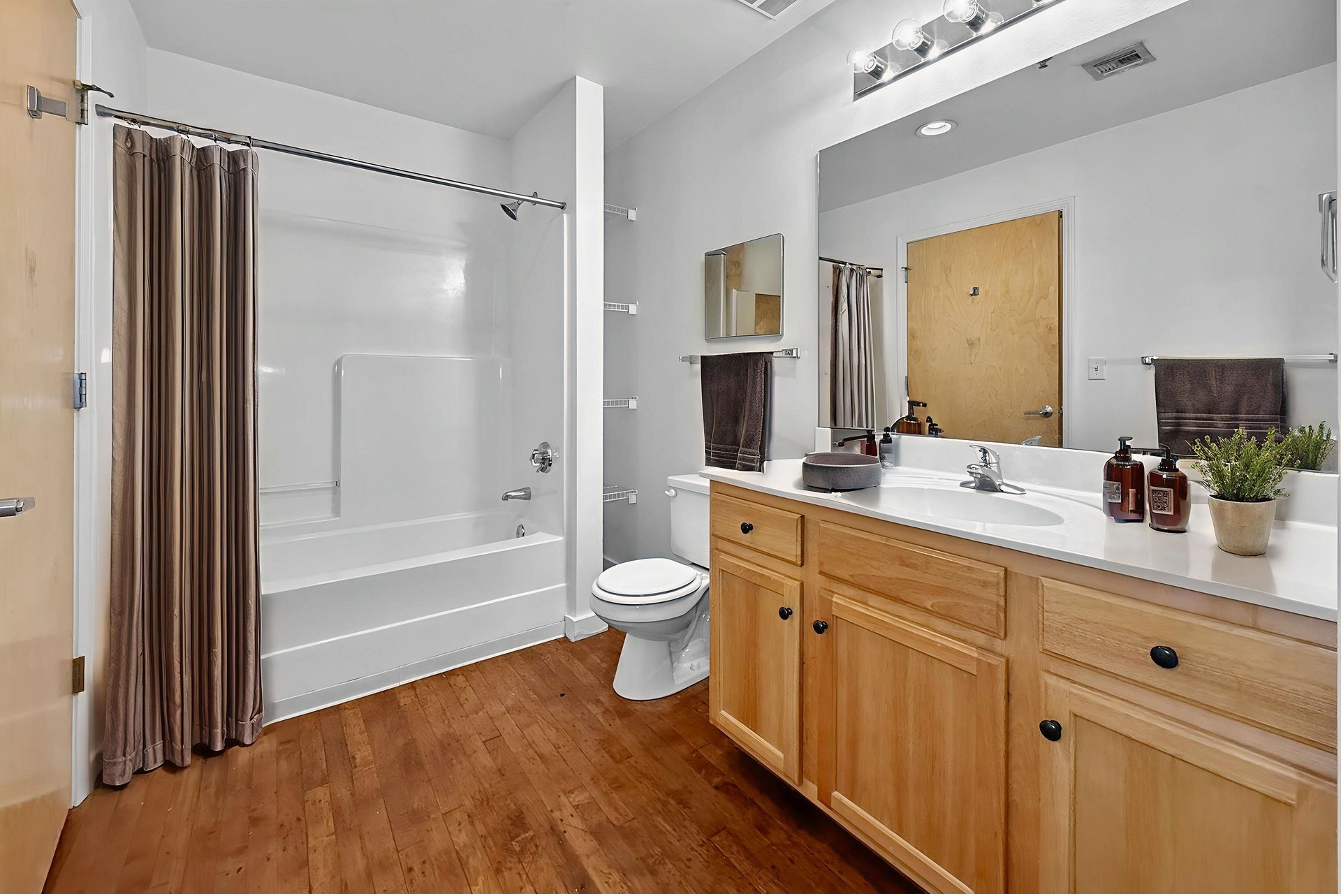 Bathroom with a bathtub, toilet, and light-wood vanity with a countertop sink, set against white walls and wood flooring.