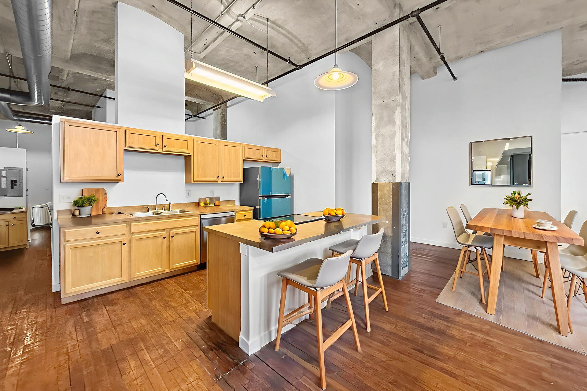 Modern kitchen featuring light wood cabinetry, a breakfast bar with two stools, and a dining area on hardwood floors.