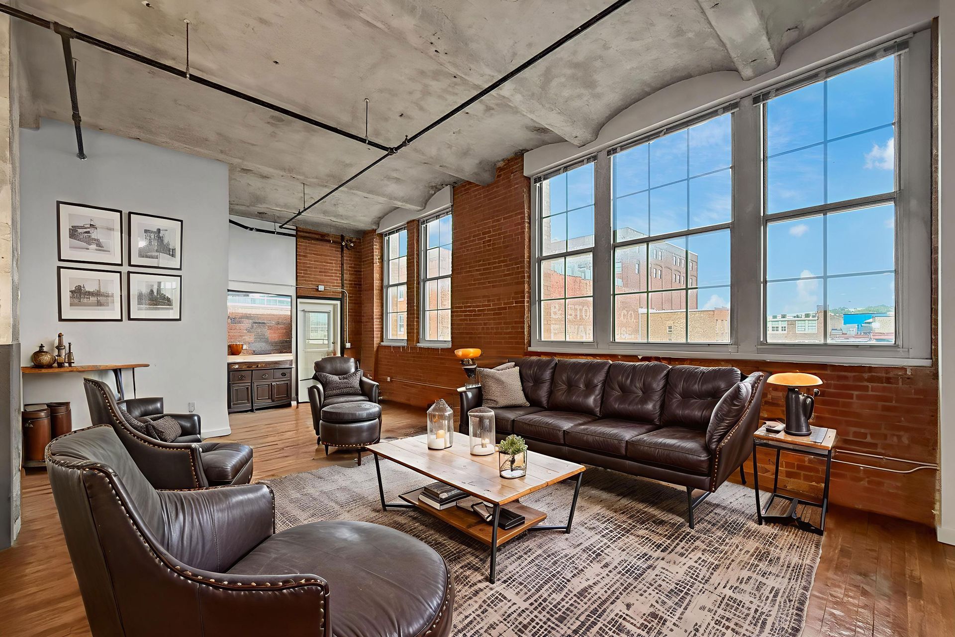 A spacious loft living room featuring exposed brick walls, a concrete ceiling, large windows, and brown leather furniture.