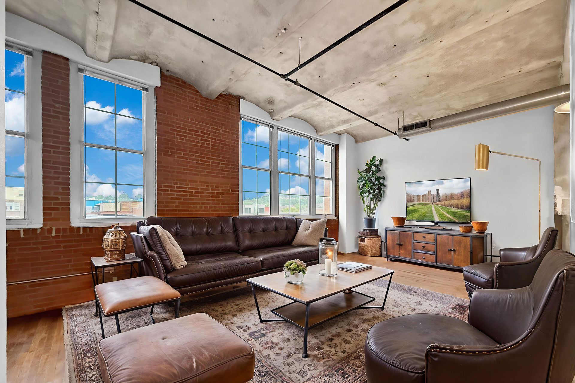 A modern loft living room with exposed brick walls, vaulted concrete ceilings, a brown leather sofa, and two armchairs.