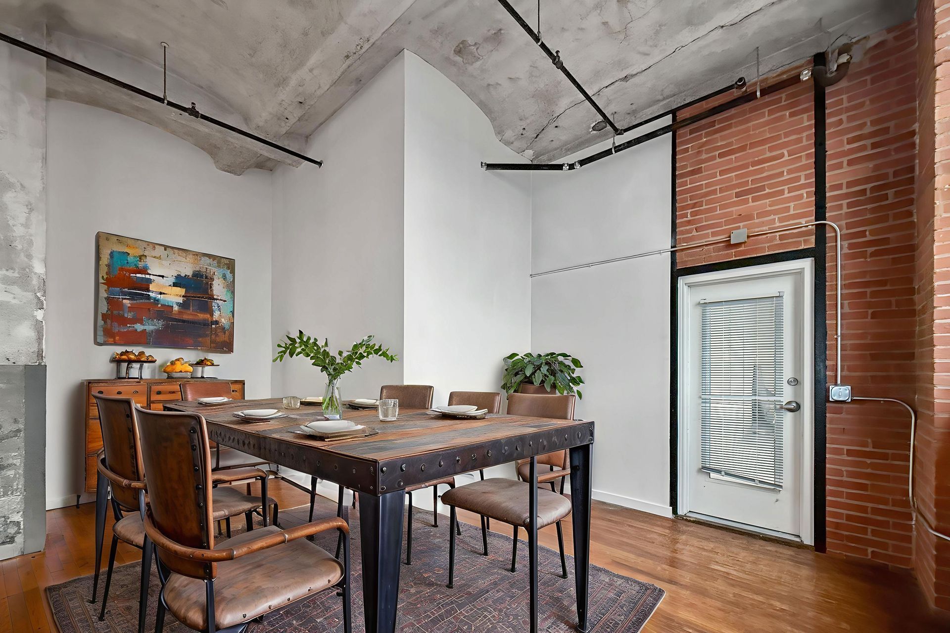 Dining area with a wooden table, six chairs, an abstract painting, and a white door, set in an industrial loft room.
