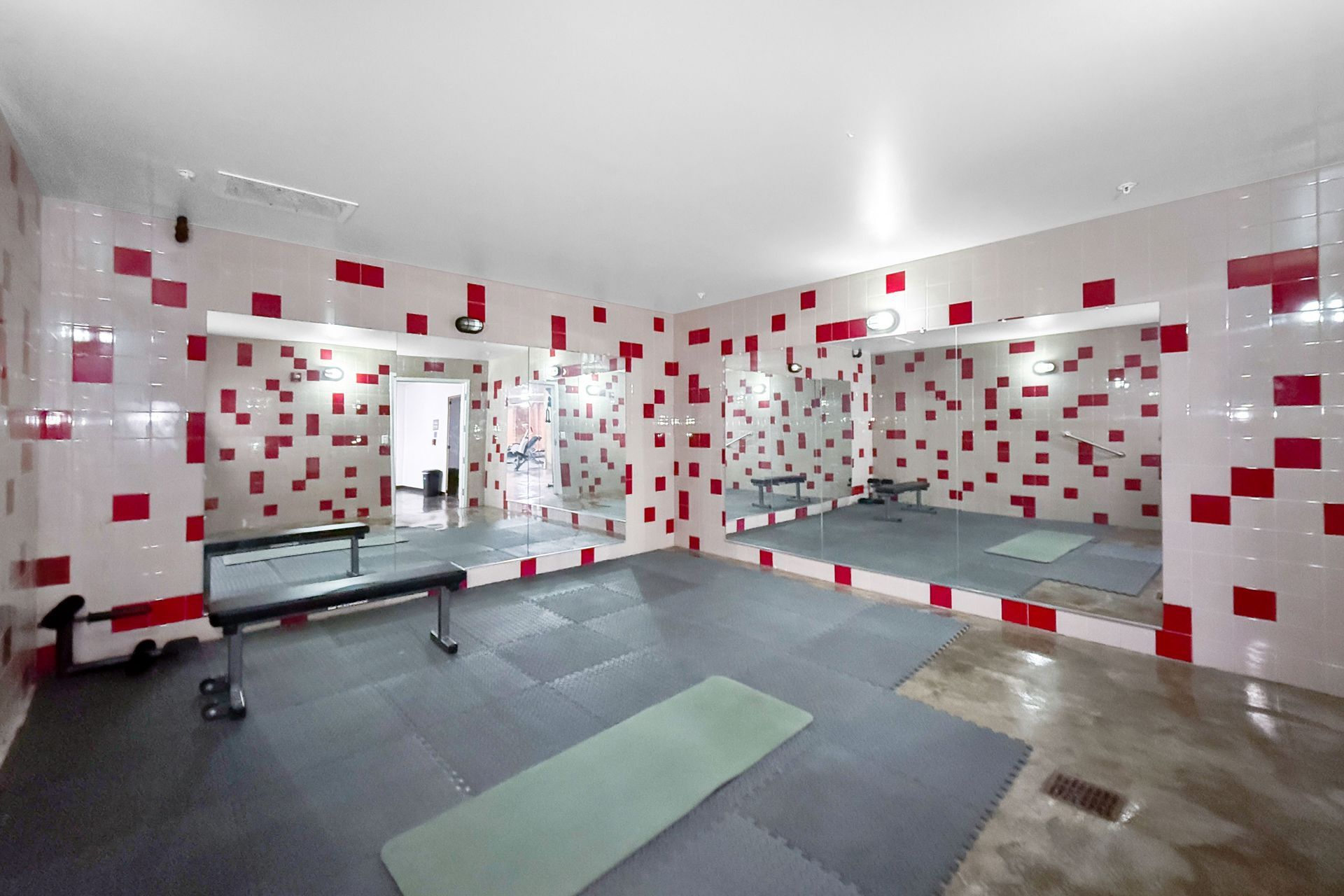 A workout room with red and white tiled walls, floor mats, a weight bench, and large wall mirrors.