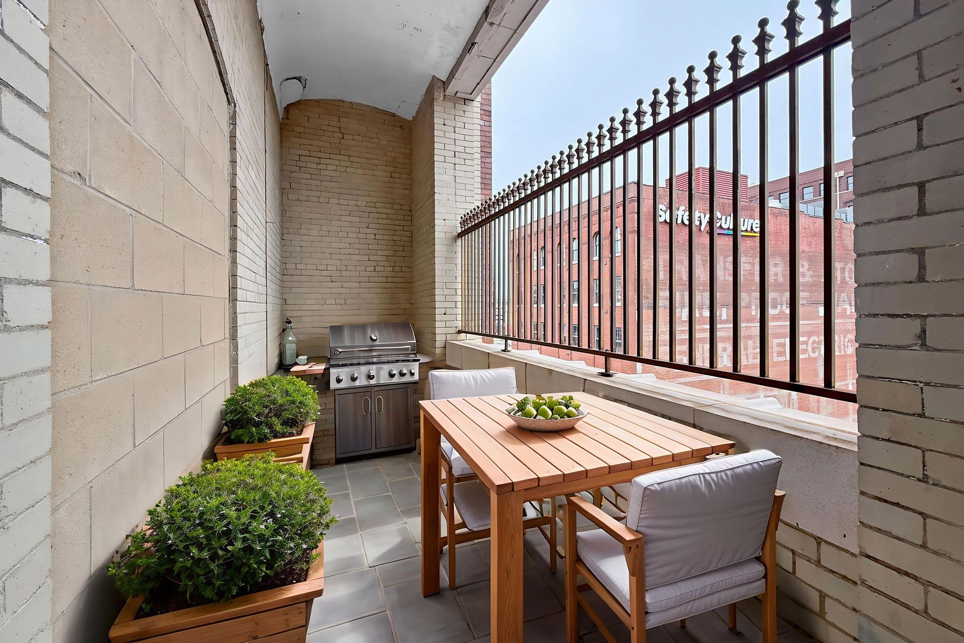 A narrow balcony with two potted shrubs, a grill, and a wooden table with two chairs.