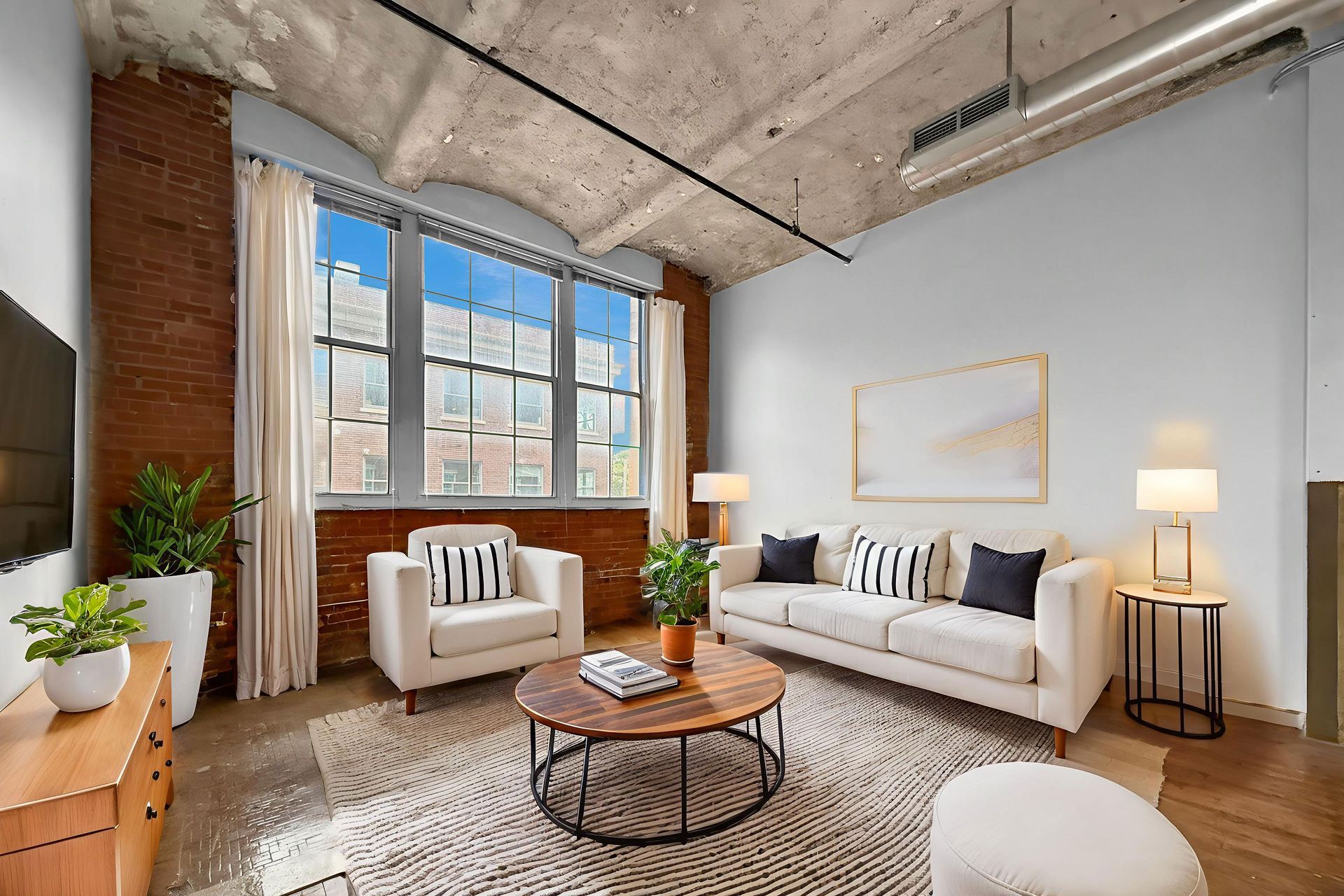 A sunlit industrial loft living room with exposed brick walls, vaulted concrete ceiling, white sofa, and armchairs.
