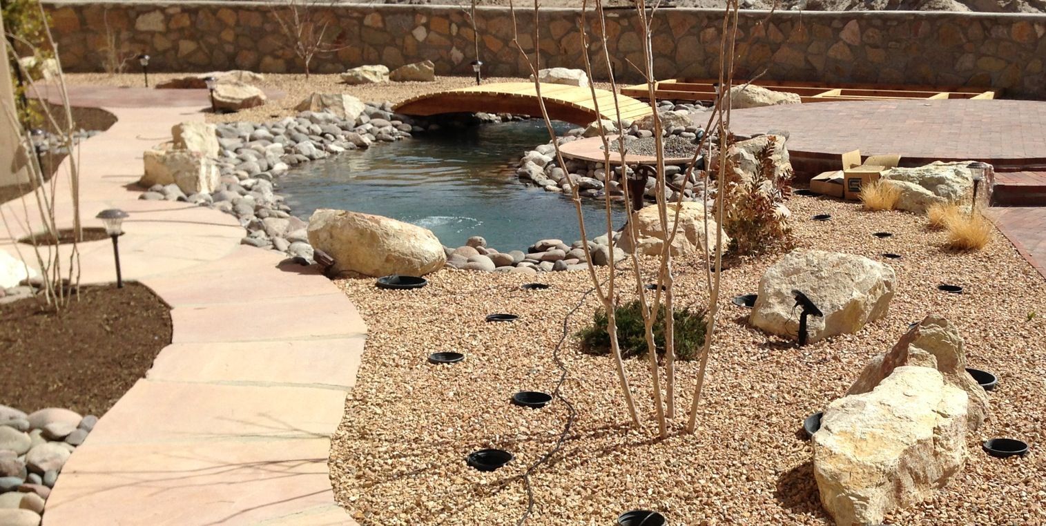 A serene backyard landscape with a pond, a small wooden bridge, and a stone pathway.