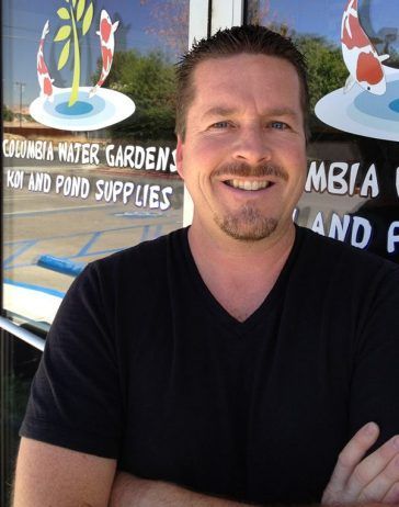 Man with arms crossed, smiling in front of a store with koi fish and water garden supplies.