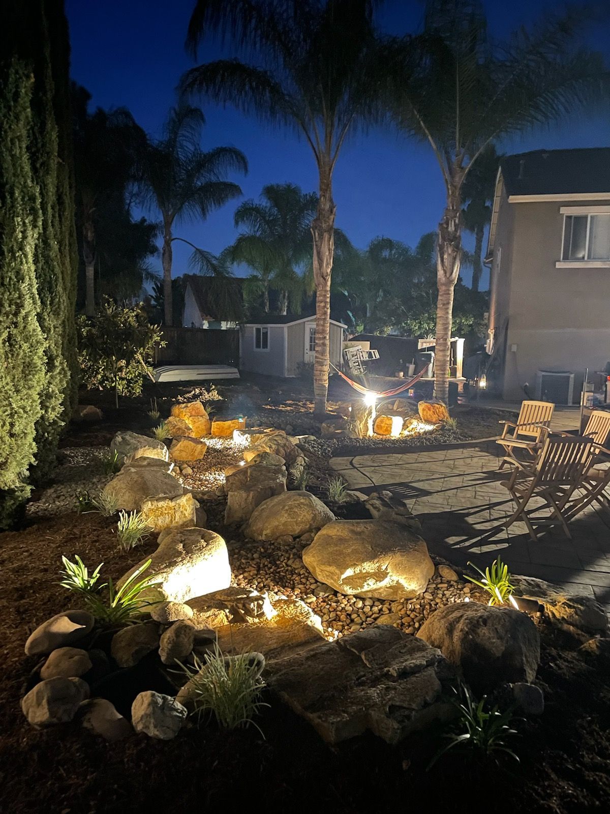 Backyard at night with illuminated rock water feature, palm trees, and home.