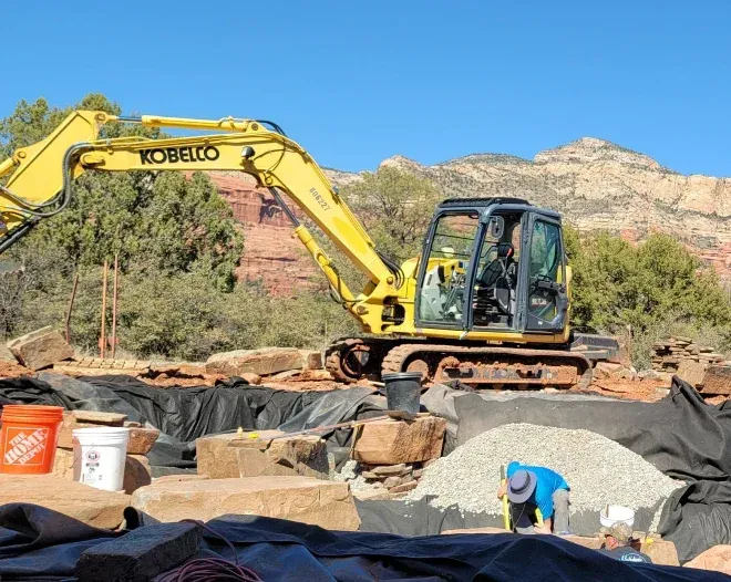 Yellow Kobelco excavator working at a construction site with rocks, gravel, and a black liner. Mountains are in the background.