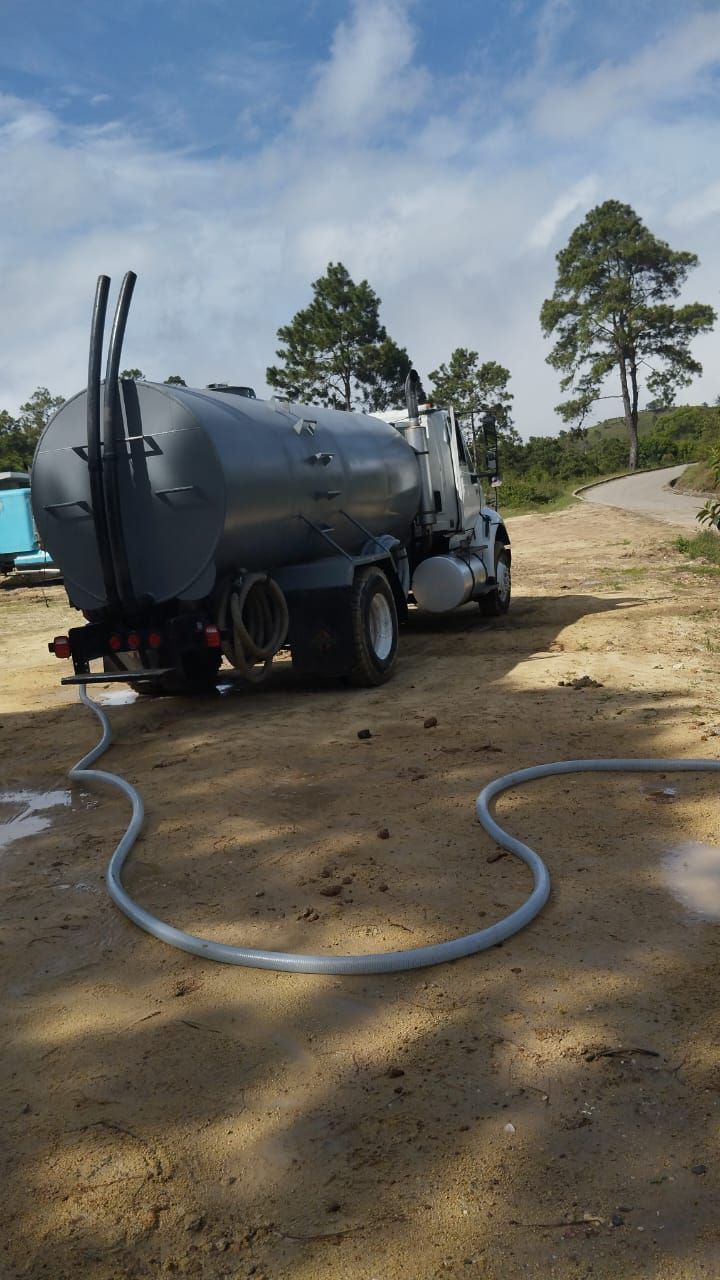 Camión cisterna gris estacionado en un camino de tierra, con la manguera extendida. Cielo azul y árboles verdes al fondo.