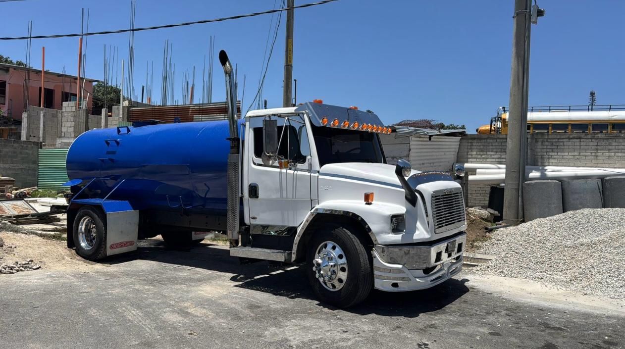 Camión cisterna blanco y azul estacionado en la calle. Luz solar brillante, una obra al fondo.