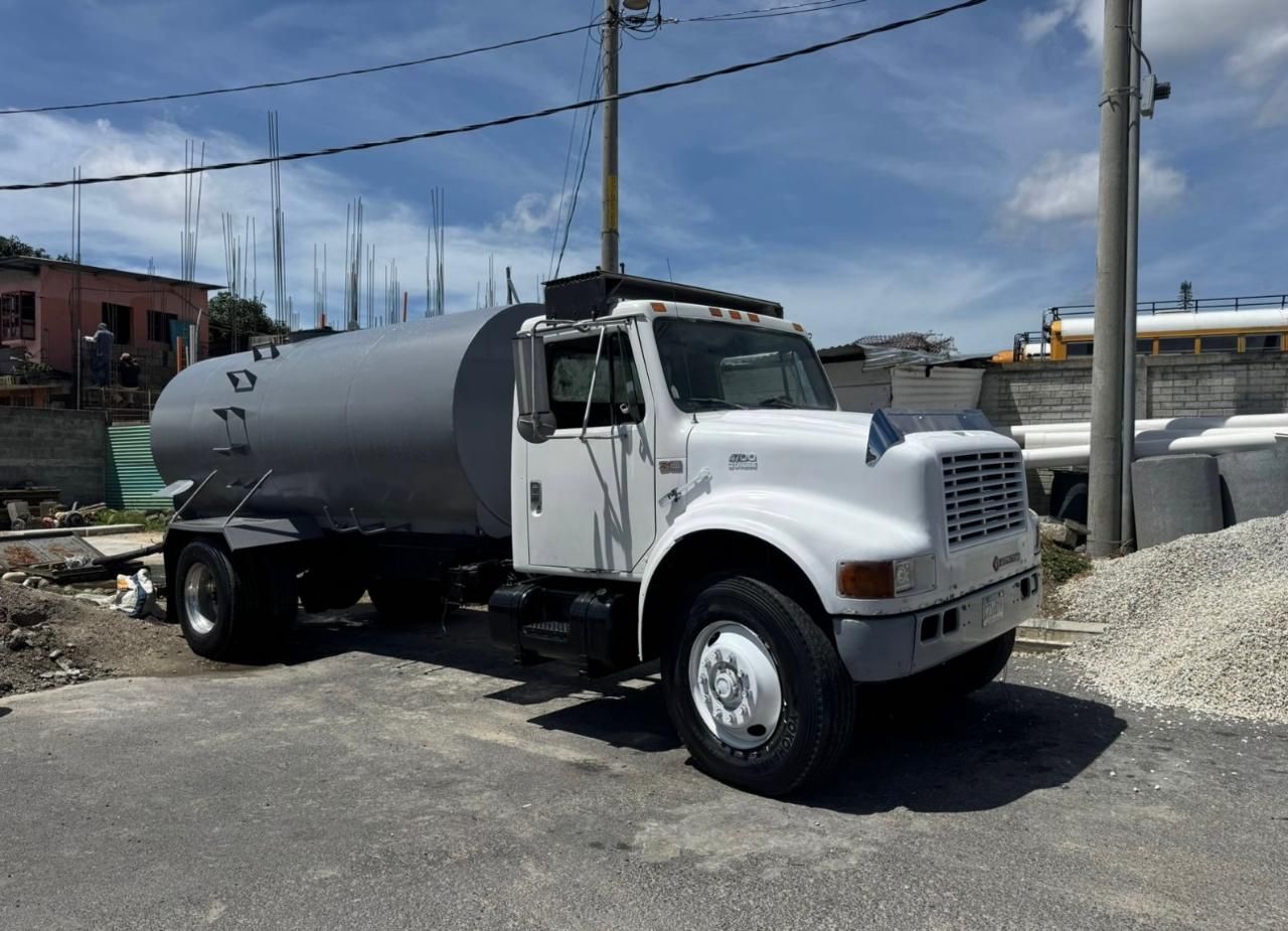 Camión cisterna de agua blanca estacionado en una carretera pavimentada, tanque gris, fondo de cielo azul.