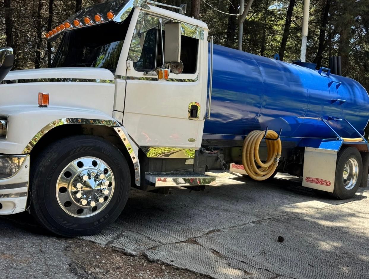 Camión cisterna de agua blanco y azul estacionado sobre una superficie pavimentada, con árboles en el fondo.