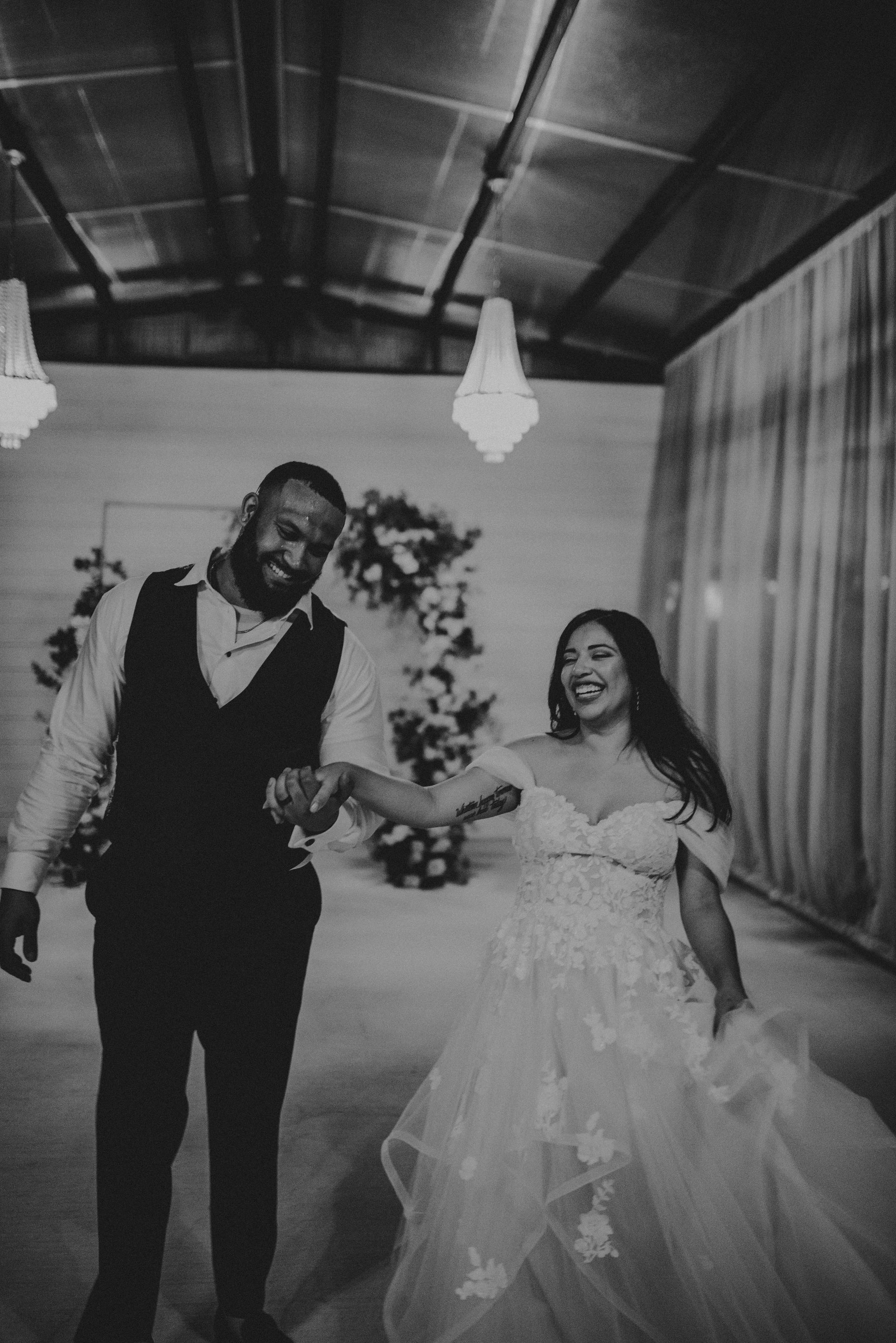 Newlywed couple smiling, holding hands, walking inside a venue. Black and white.