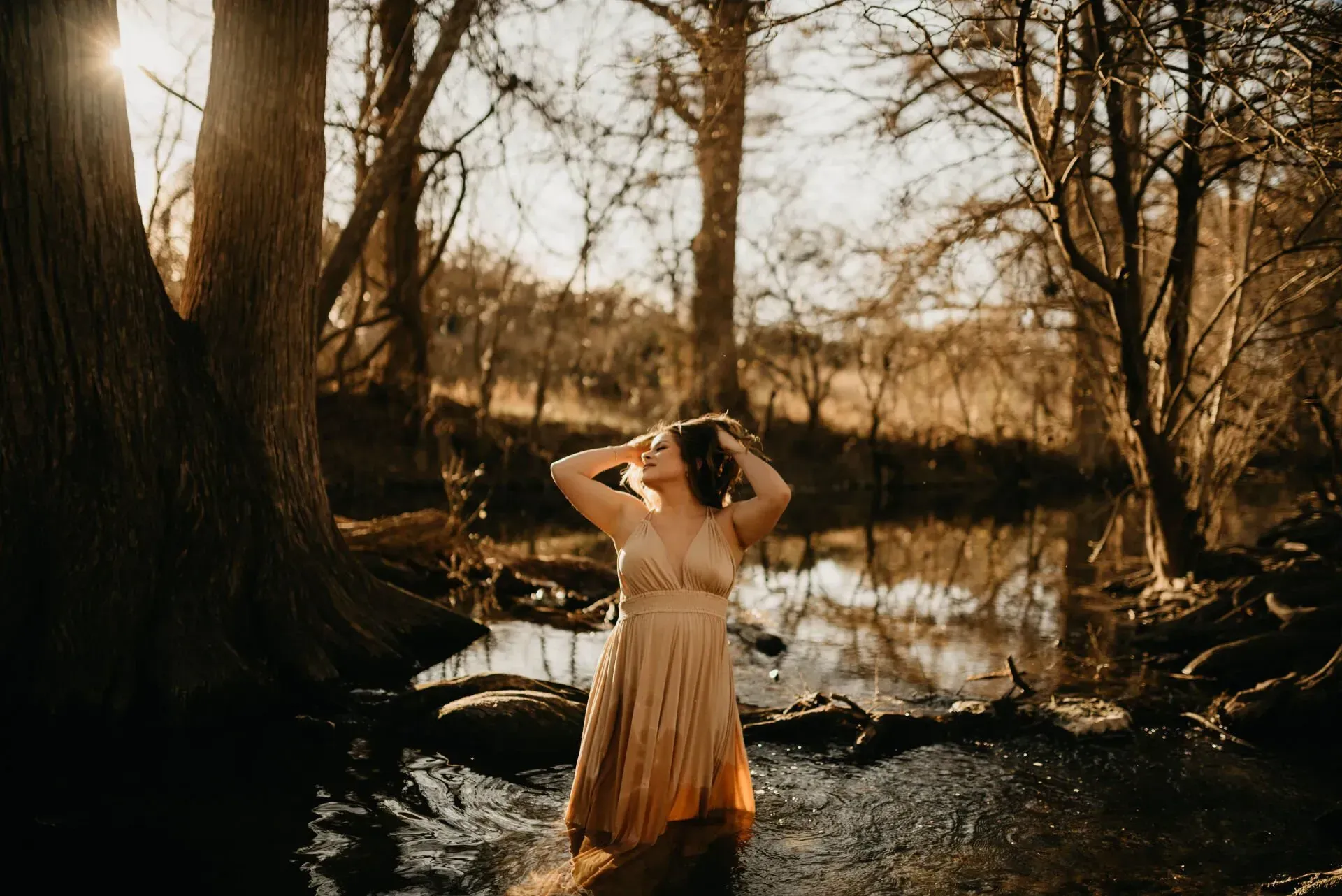 Woman in gold dress stands in stream with arms raised, backlit by the sun, surrounded by trees.