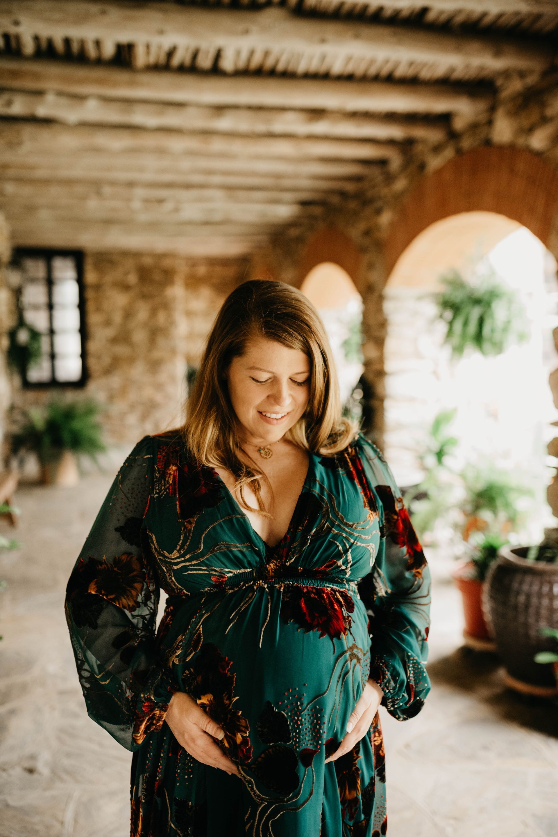Pregnant woman in green dress, hands on her belly, smiles in a stone archway.