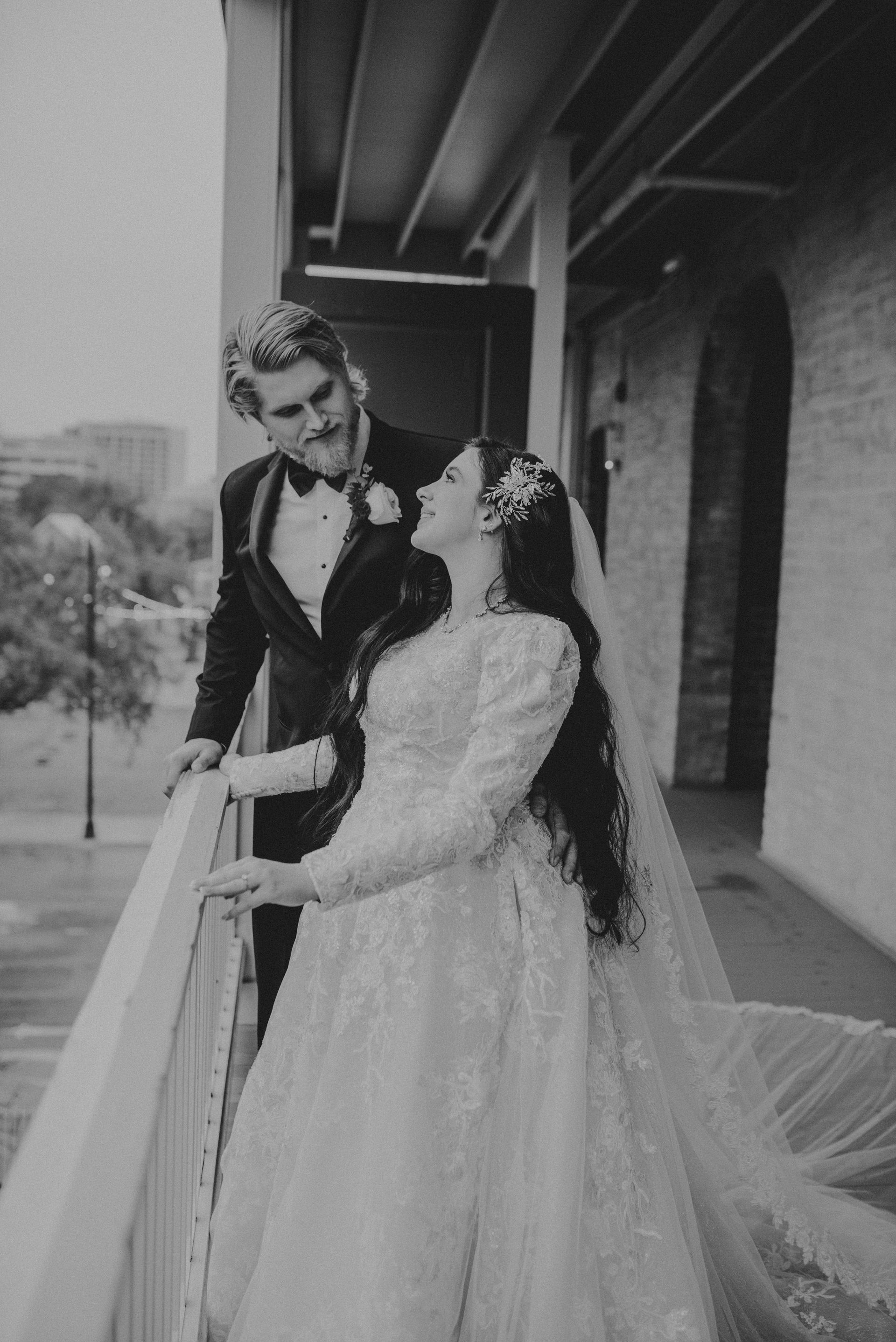 Bride in white gown gazes up at groom in tuxedo on balcony. Black and white photo.