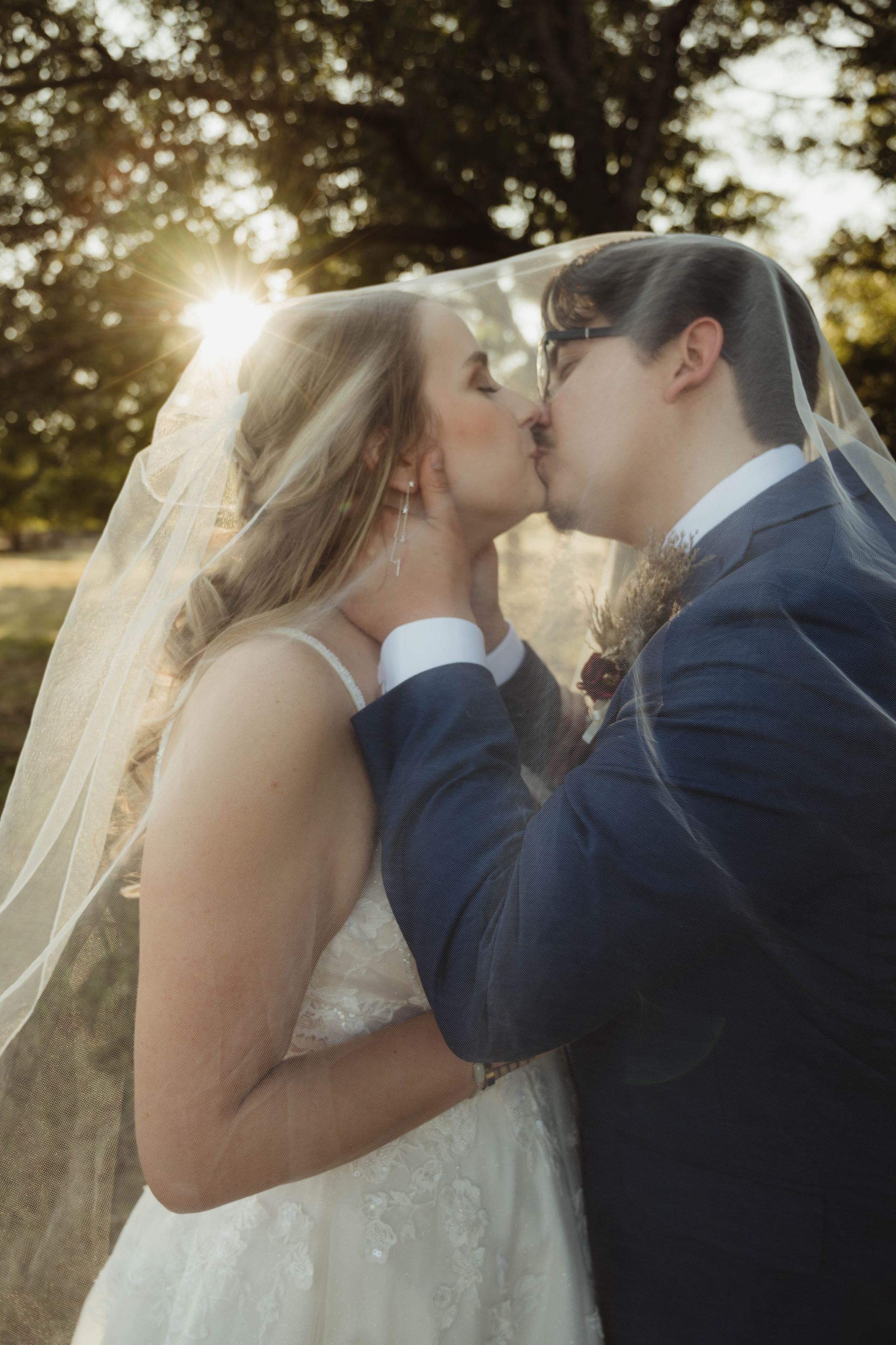 Bride and groom kissing under a veil, sun shining behind them.