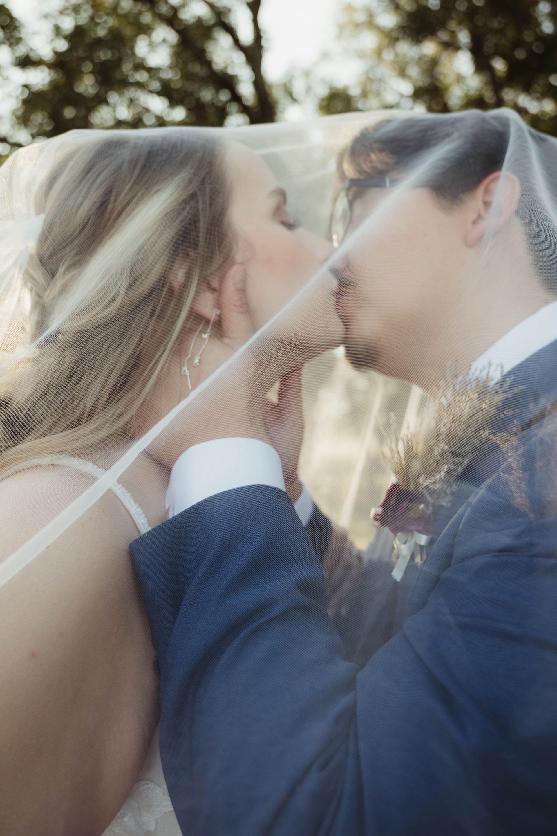 Bride and groom kissing under a sheer veil; the man in a blue suit, outdoor setting.