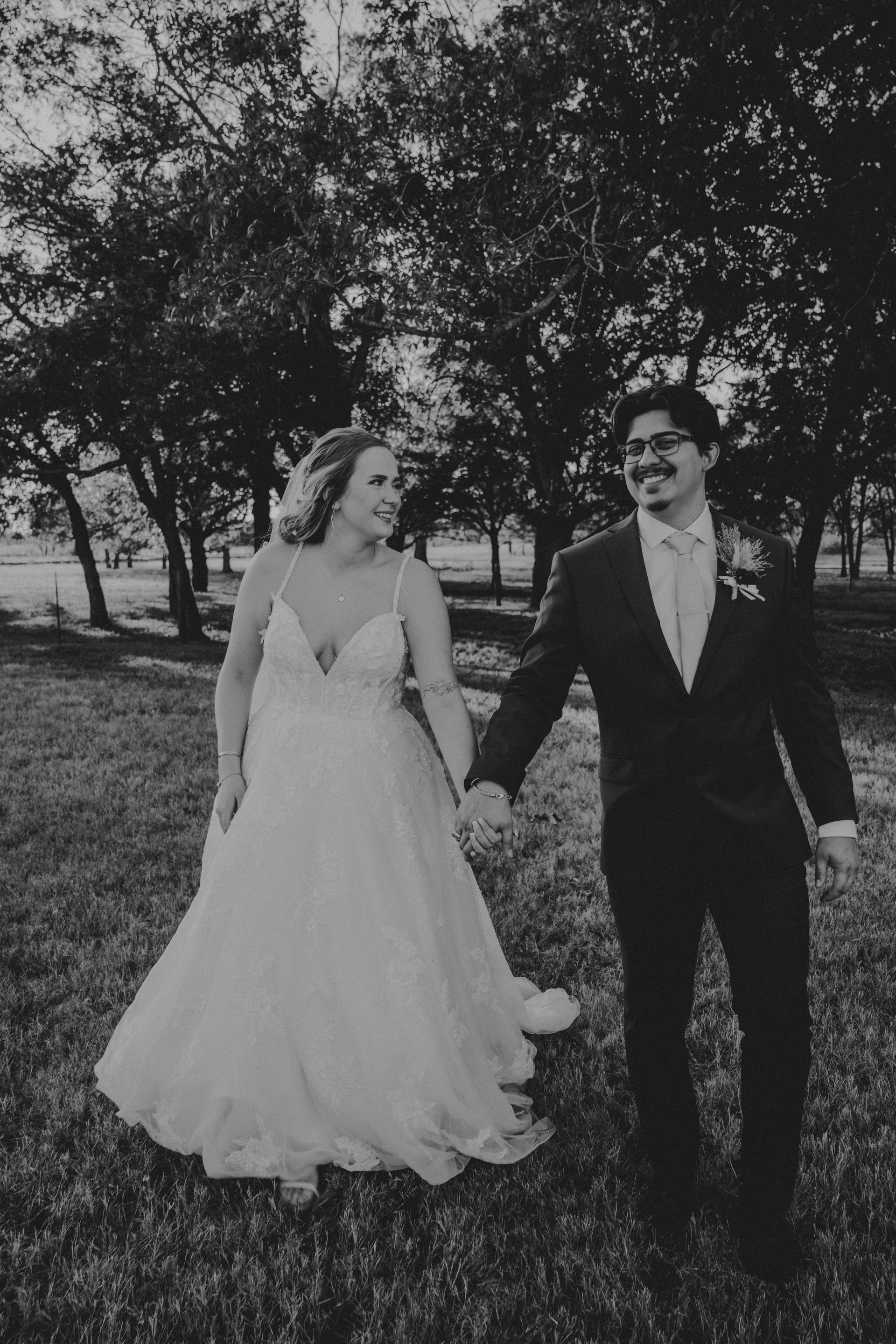 Newlyweds holding hands, smiling, in a grassy area with trees, in black and white.