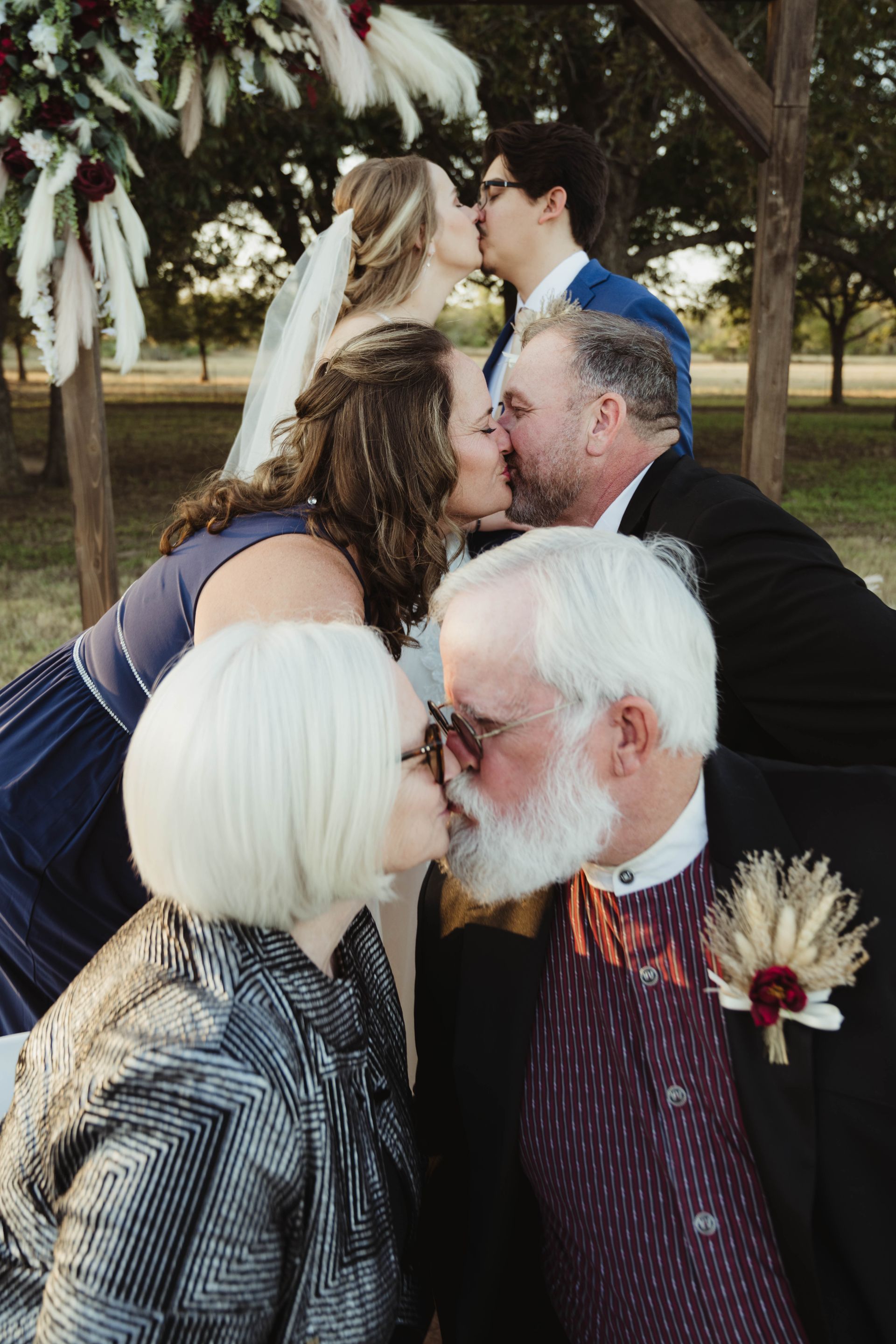Wedding group kissing under an arch. Couples kiss, dressed in wedding attire. Outdoor setting, neutral tones.