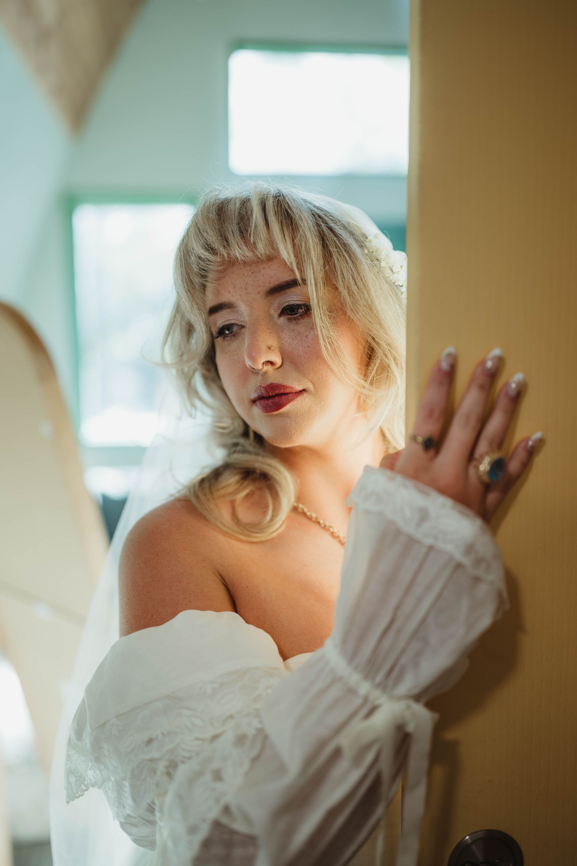 Wedding Bride in white dress, leaning against a yellow wall, looking thoughtful. Sunlight, ornate sleeves, jewelry visible.