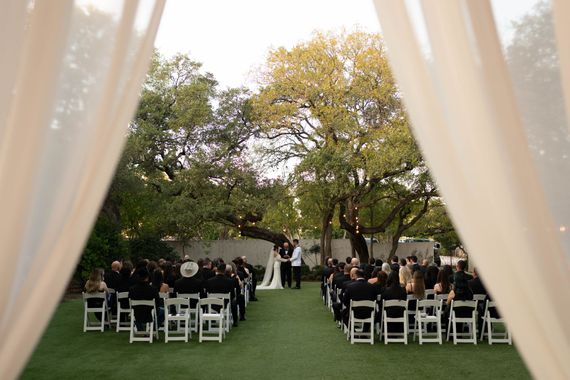Wedding ceremony outdoors, couple at altar, guests seated on white chairs, seen through sheer curtains.