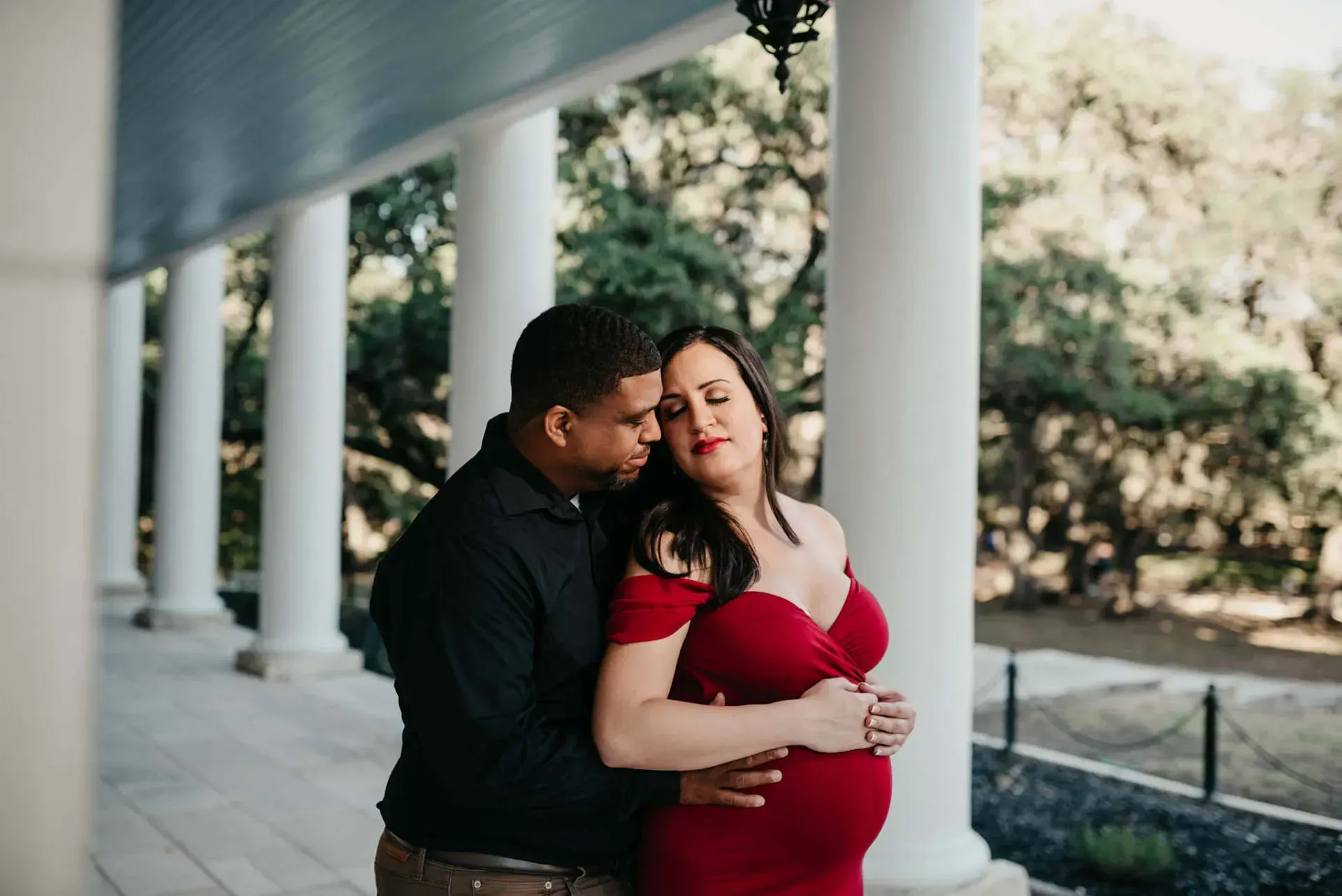 Pregnant woman in red dress embraced by man in black shirt, outdoors near white columns.