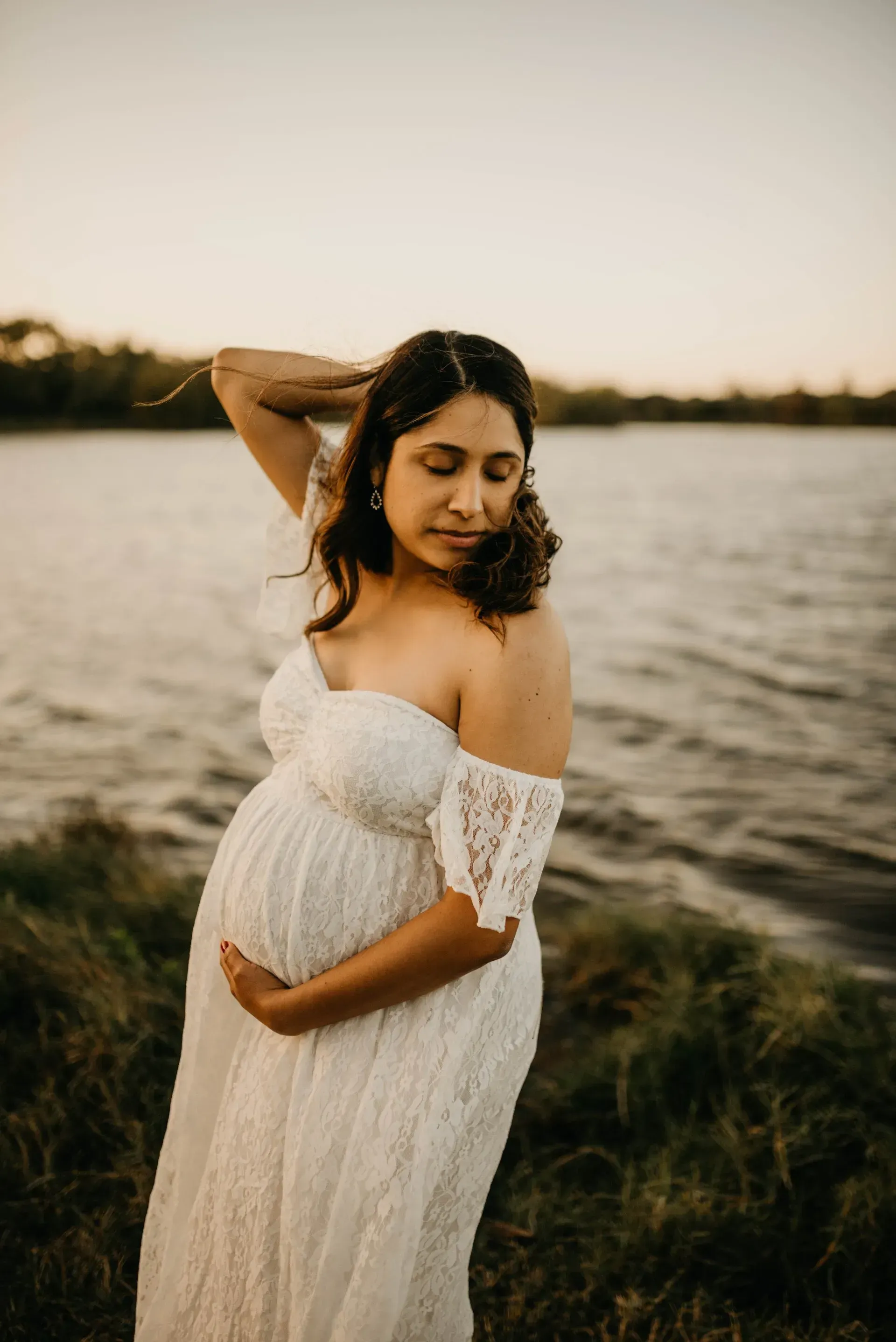 Pregnant woman in a white lace dress by a lake, holding belly, arm in hair, eyes closed.
