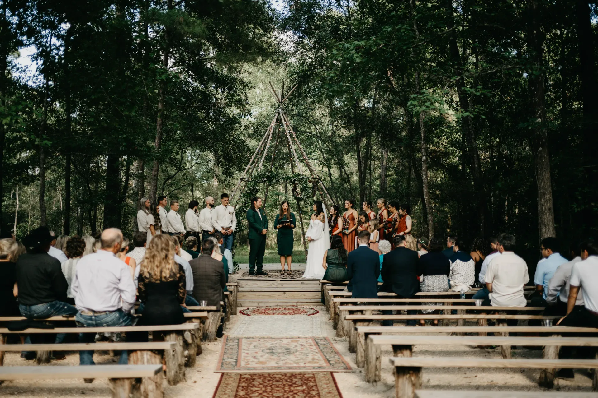 Wedding ceremony in a forest clearing. Guests seated on benches face the couple and wedding party under a teepee.