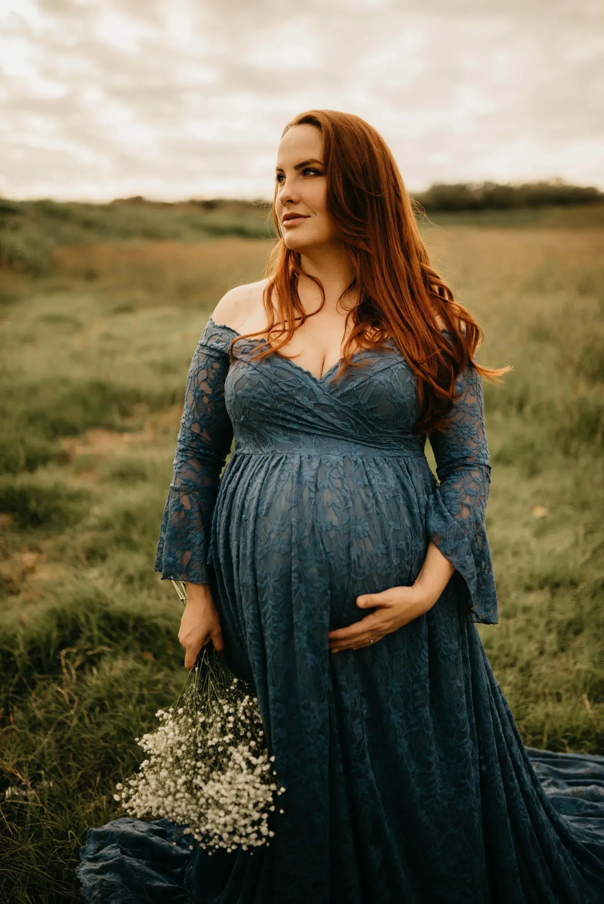 Pregnant woman with long red hair in a blue lace gown, holding flowers, in a field, looking to the side.