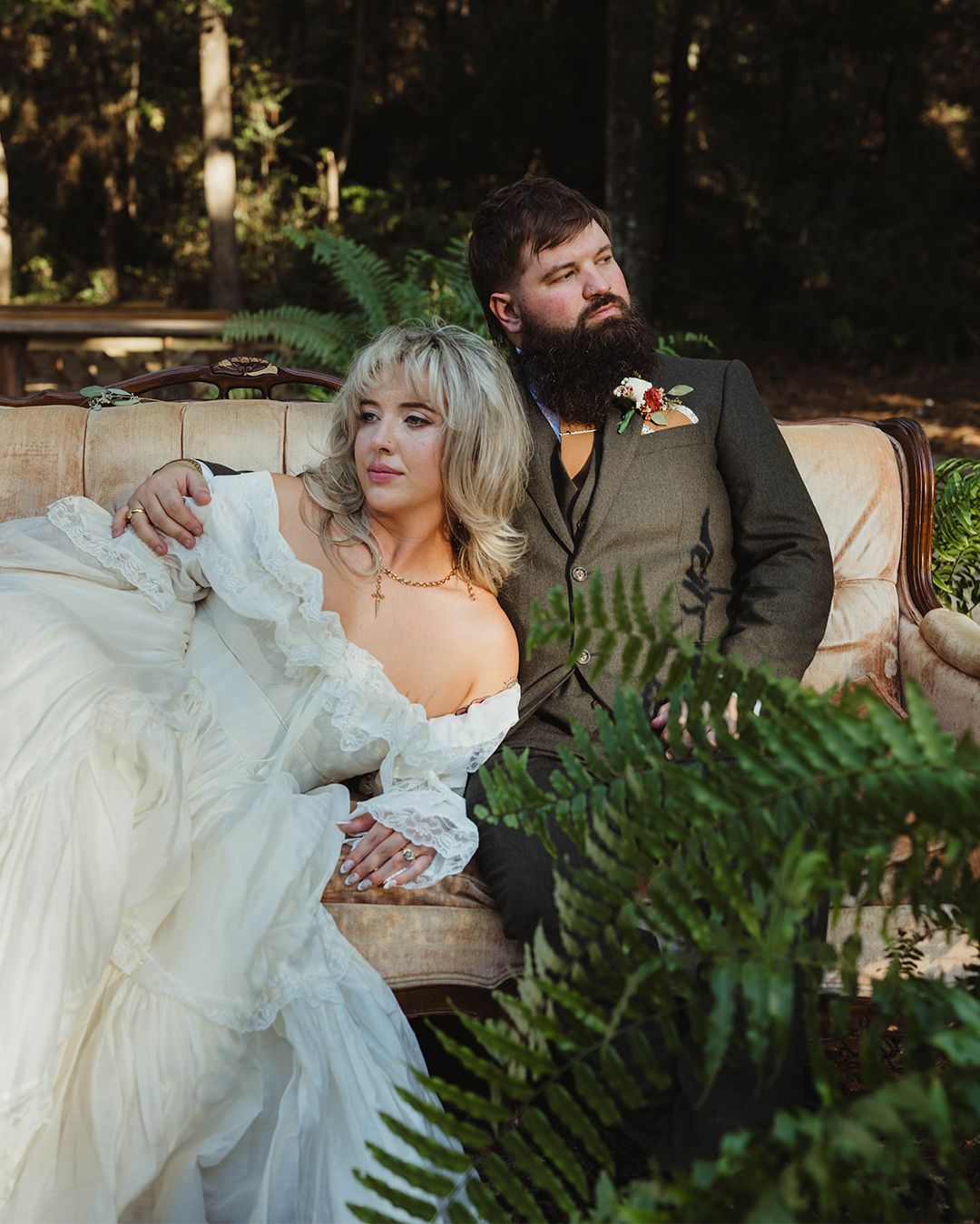 Bride and groom on a vintage couch in a forest. She wears a white gown. He has a beard and tweed jacket.