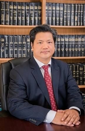 A man in a suit and tie is sitting at a desk in front of a bookshelf.