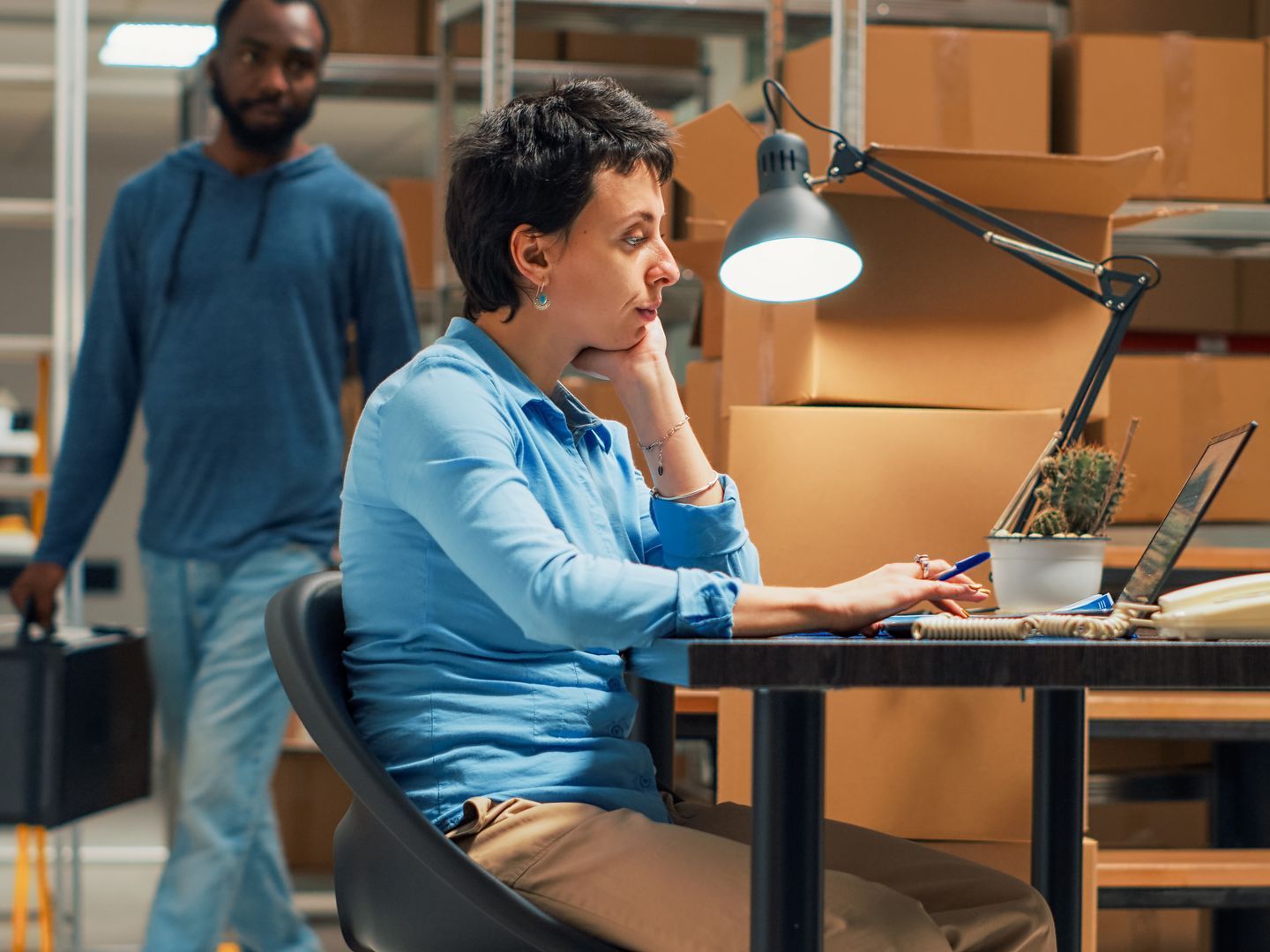 A man is walking behind a woman sitting at a desk using a laptop computer.