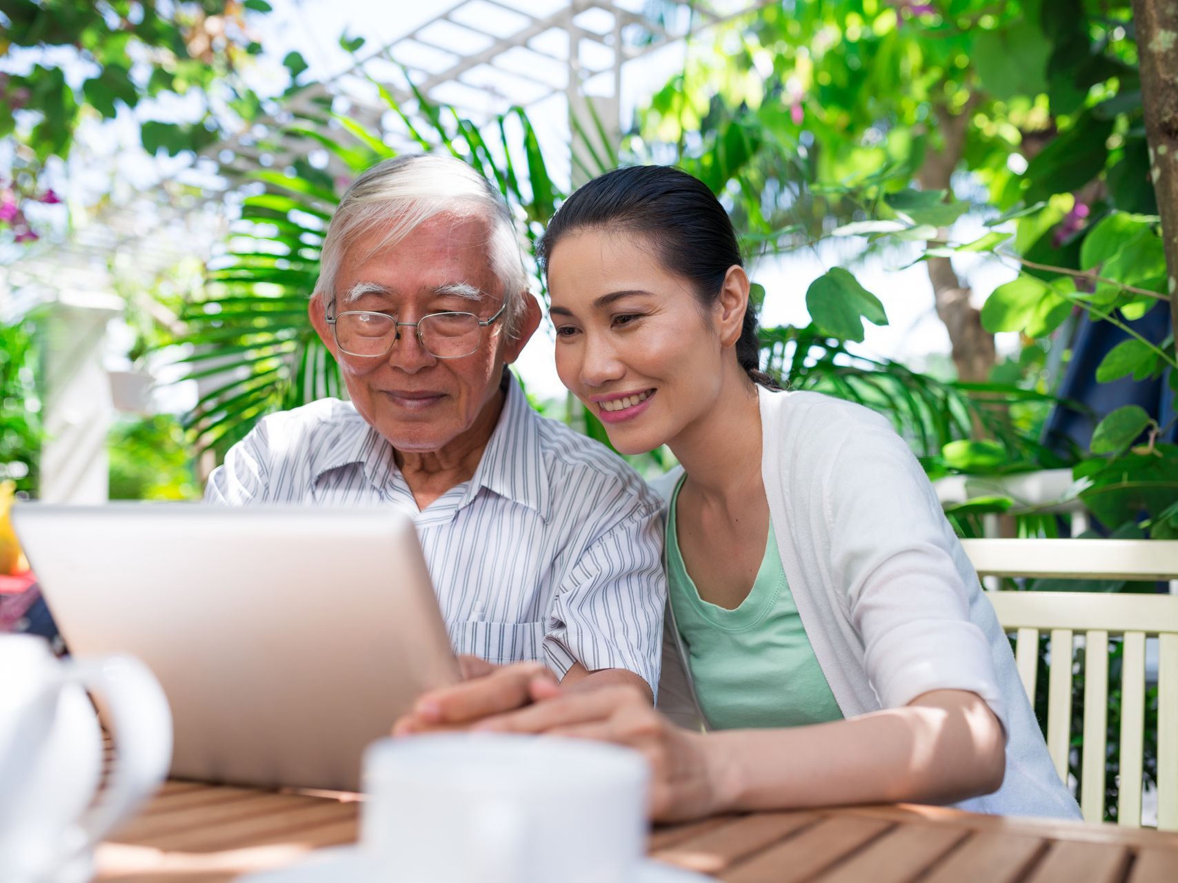 An elderly man and a young woman are sitting at a table looking at a tablet computer.