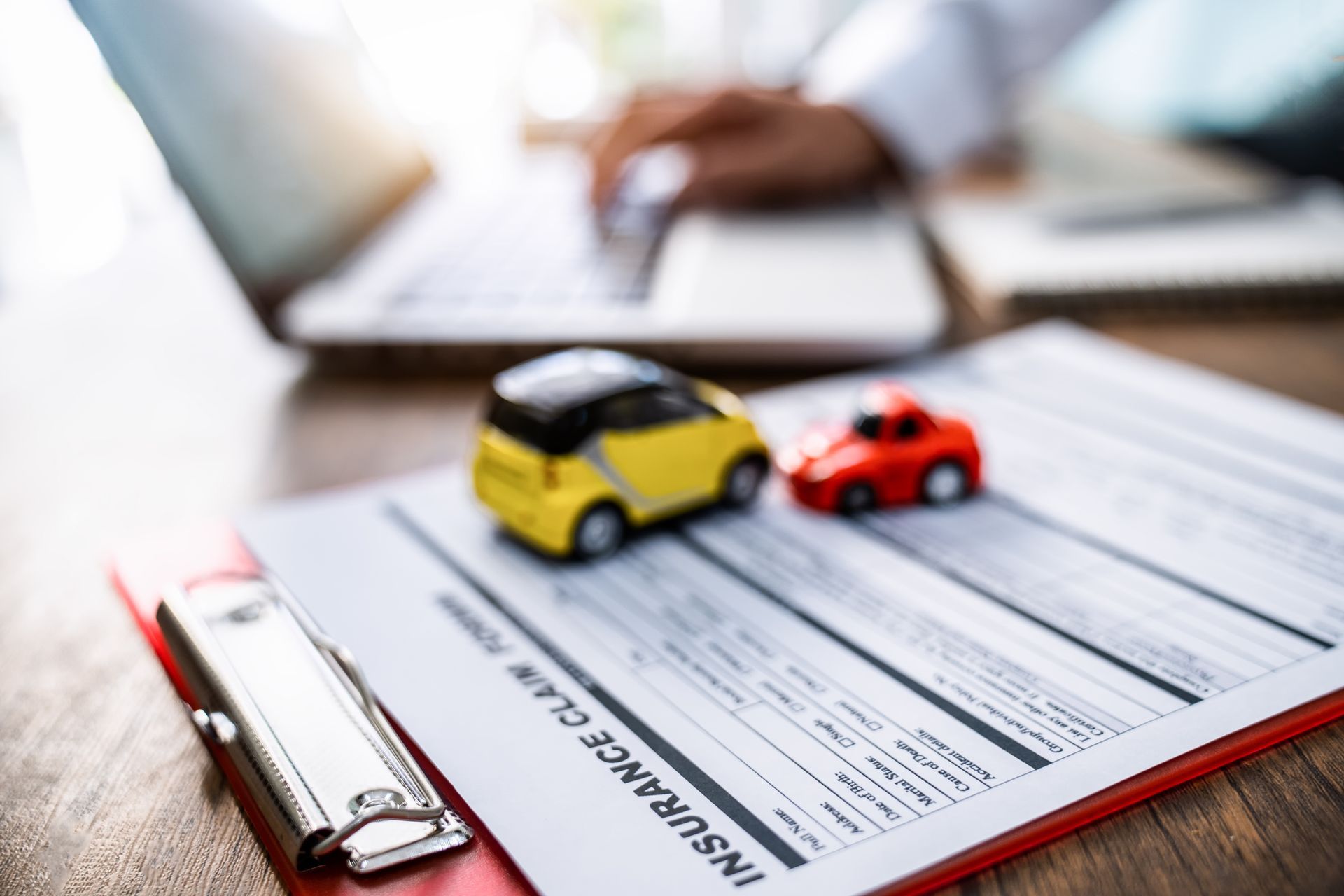 An insurance claim form with two toy cars on top. An attorney on a laptop is in the background.