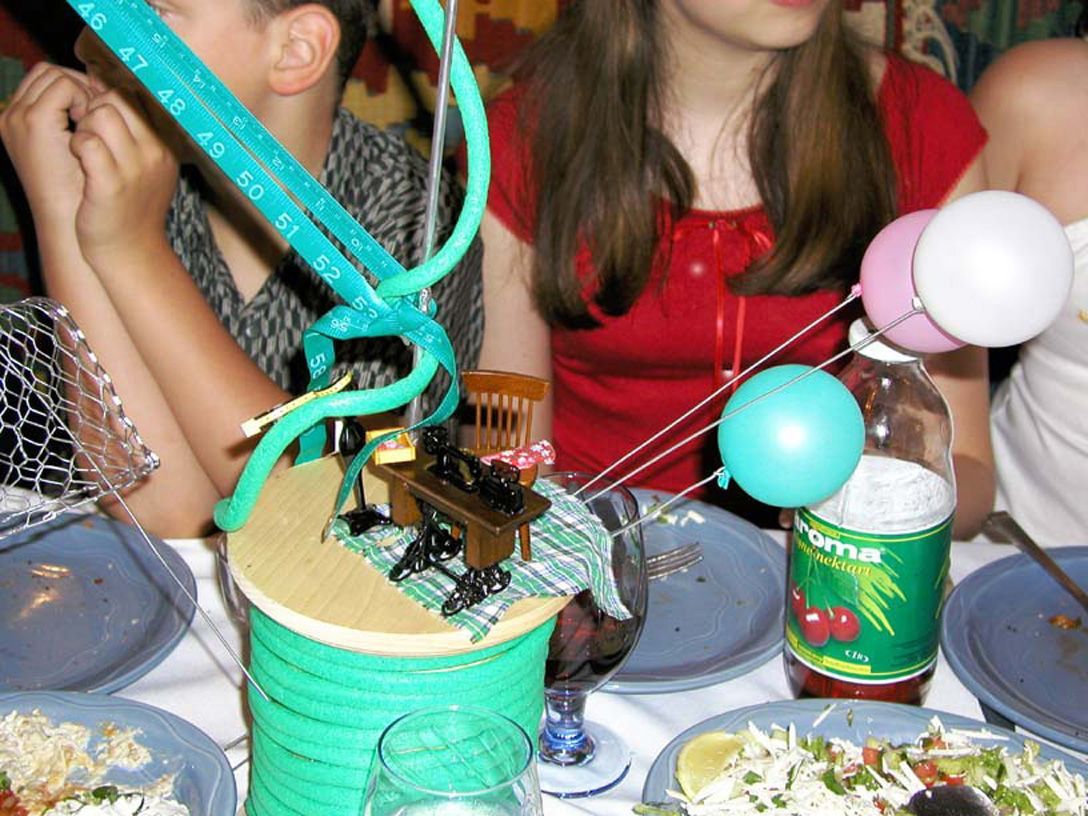 A table centerpiece with a toy piano, balloons, and drink bottle; people are seated around the table.