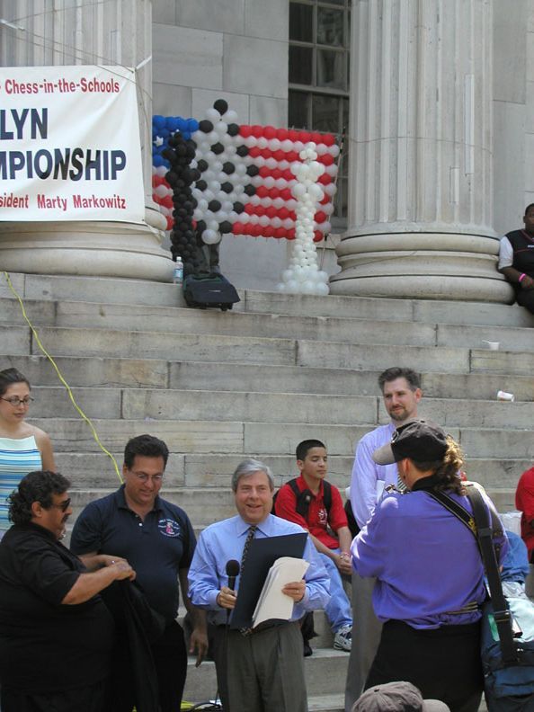 People at a Brooklyn chess championship with an American flag balloon display and speaker.