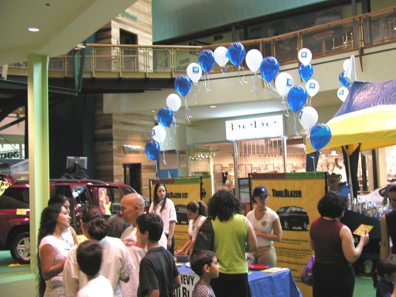 People at event in shopping mall, blue/white balloon arch, booths set up with displays, red vehicle visible.