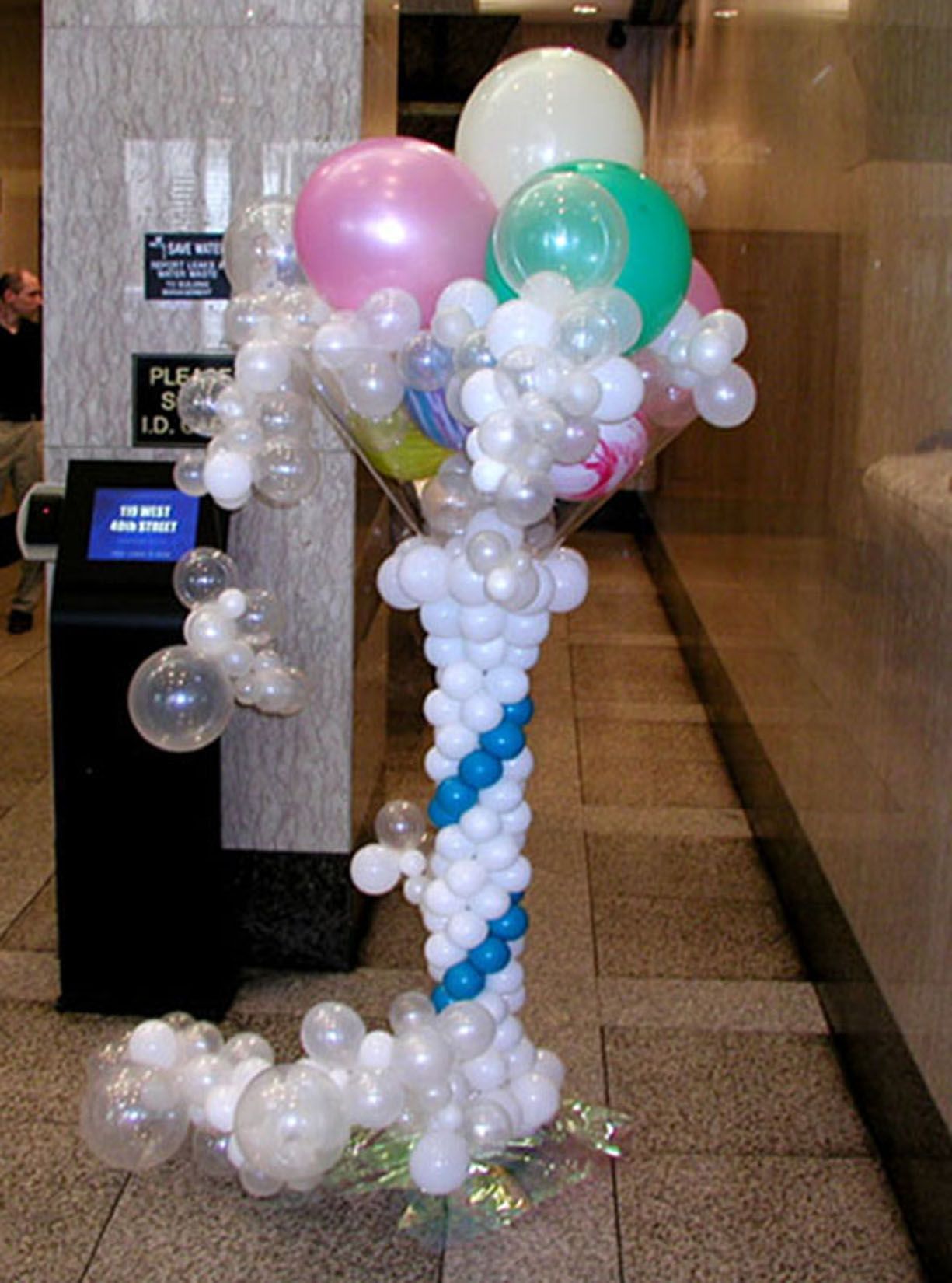 Balloon sculpture in a building lobby; white and pastel balloons in a spiral column and bouquet arrangement.