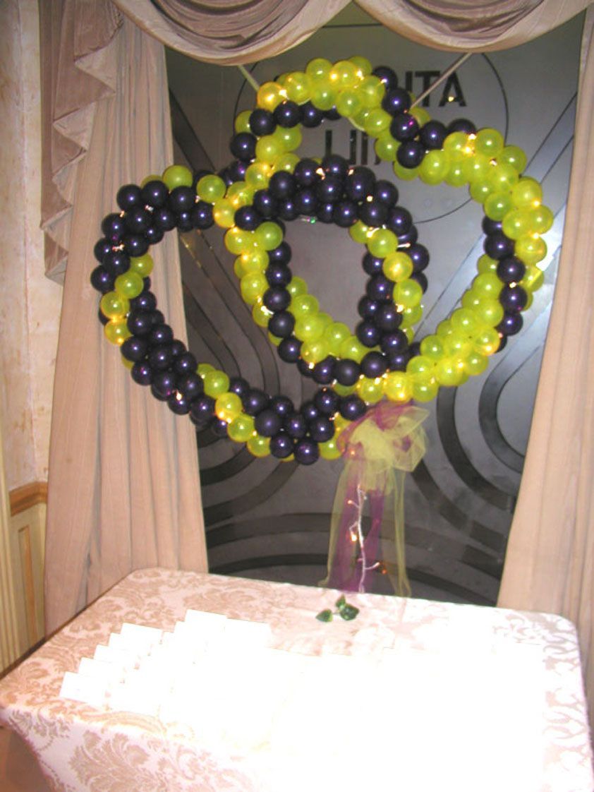 Two interlocked heart-shaped balloon decorations, alternating lime green and dark purple, above a table with a floral arrangement.