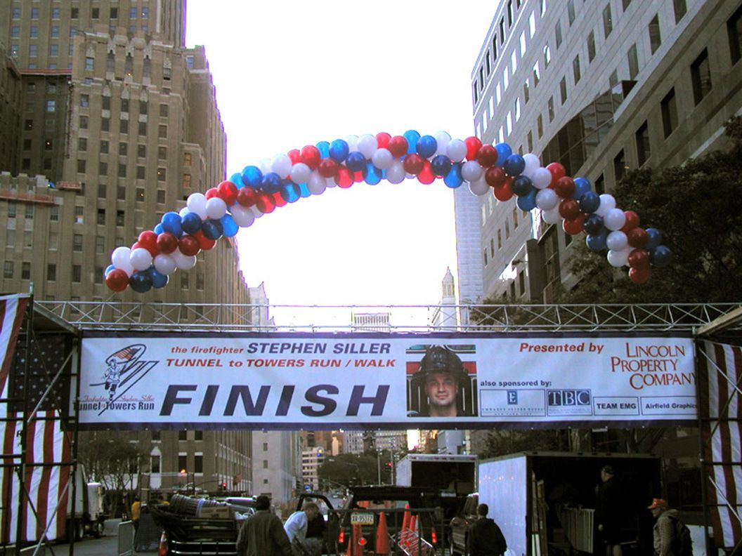 Finish line banner with red, white, and blue balloon arch. People gather in front of skyscrapers.