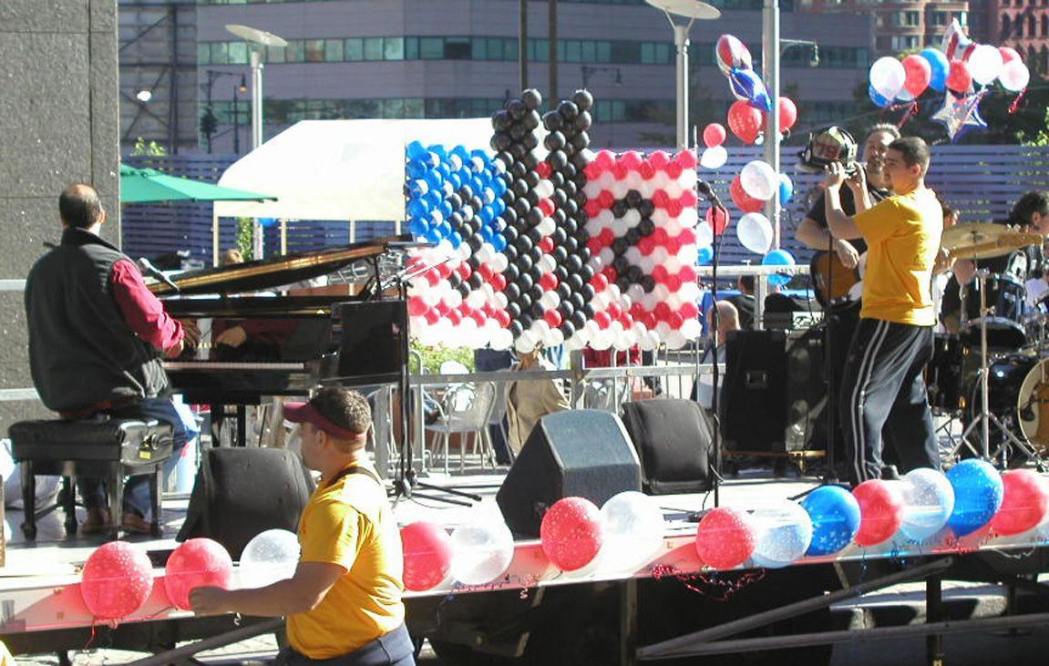 Band playing on outdoor stage decorated with red, white, and blue balloons. A piano player and other musicians perform.