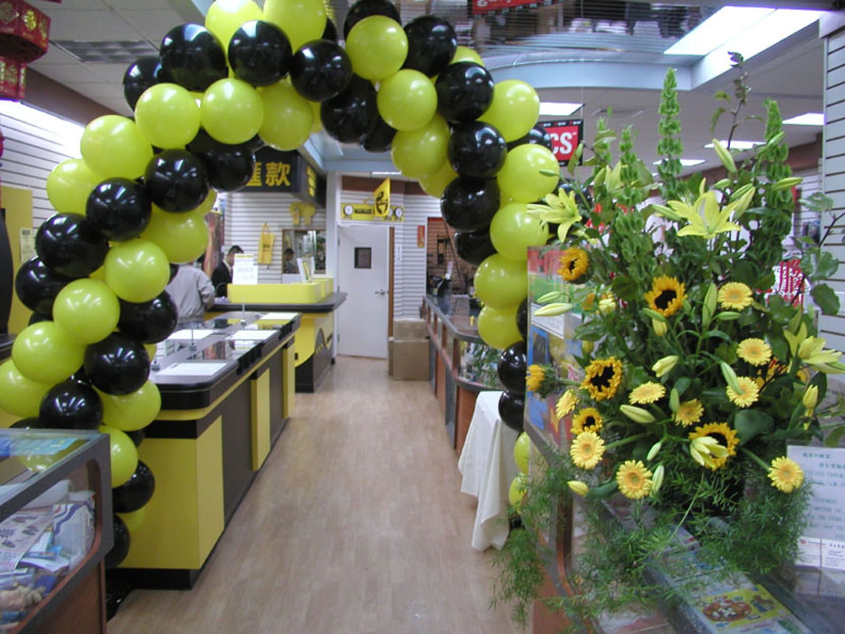 Balloon arch of yellow and black balloons at the entrance of a store, next to a flower arrangement.